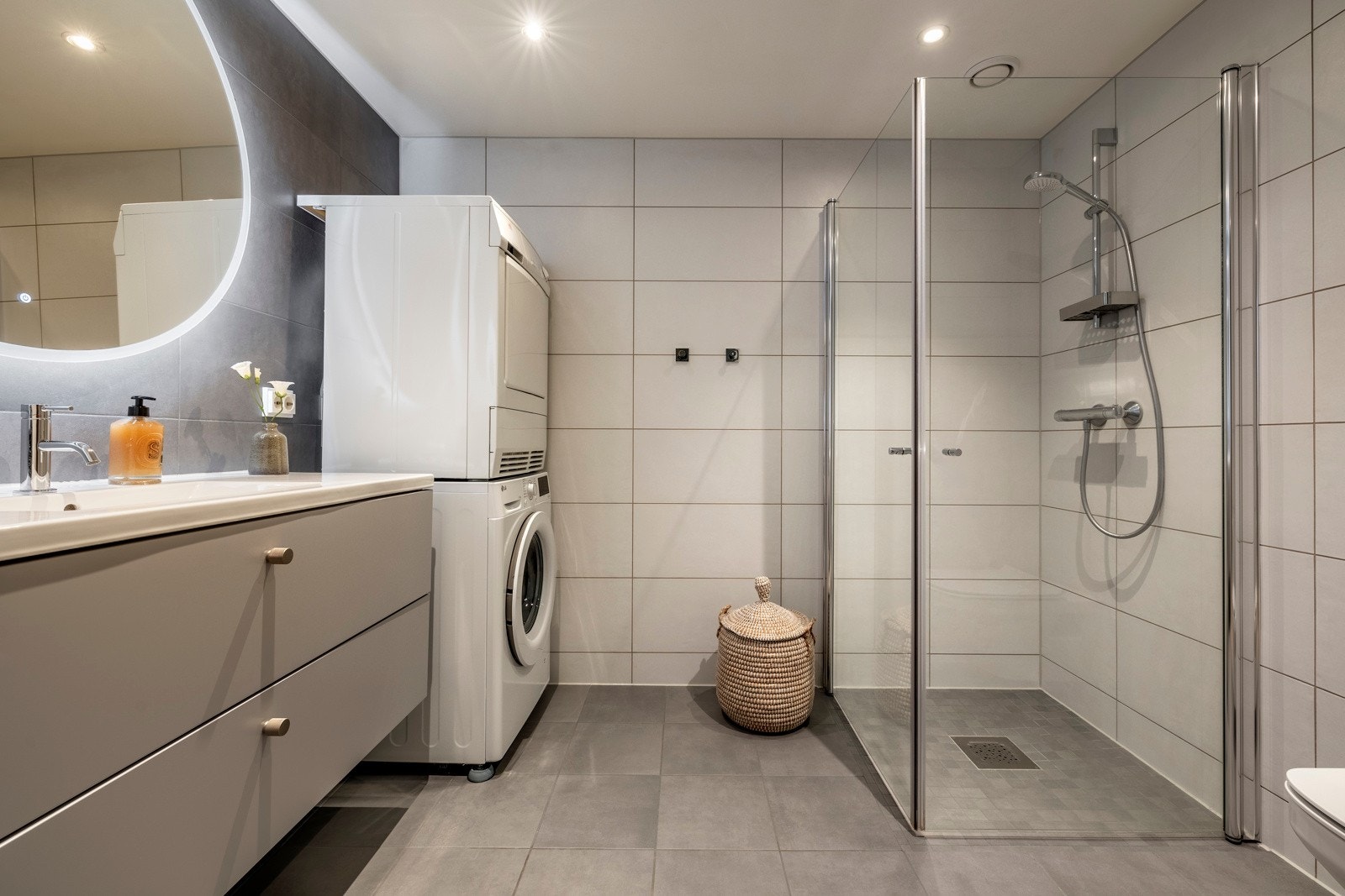 Baderomsinnredningen er i lys grå, glatt utførelse og består av en servant nedfelt i benkeplate, en praktisk skuffeseksjon og et rundt speil med integrert belysning. Galleribilde