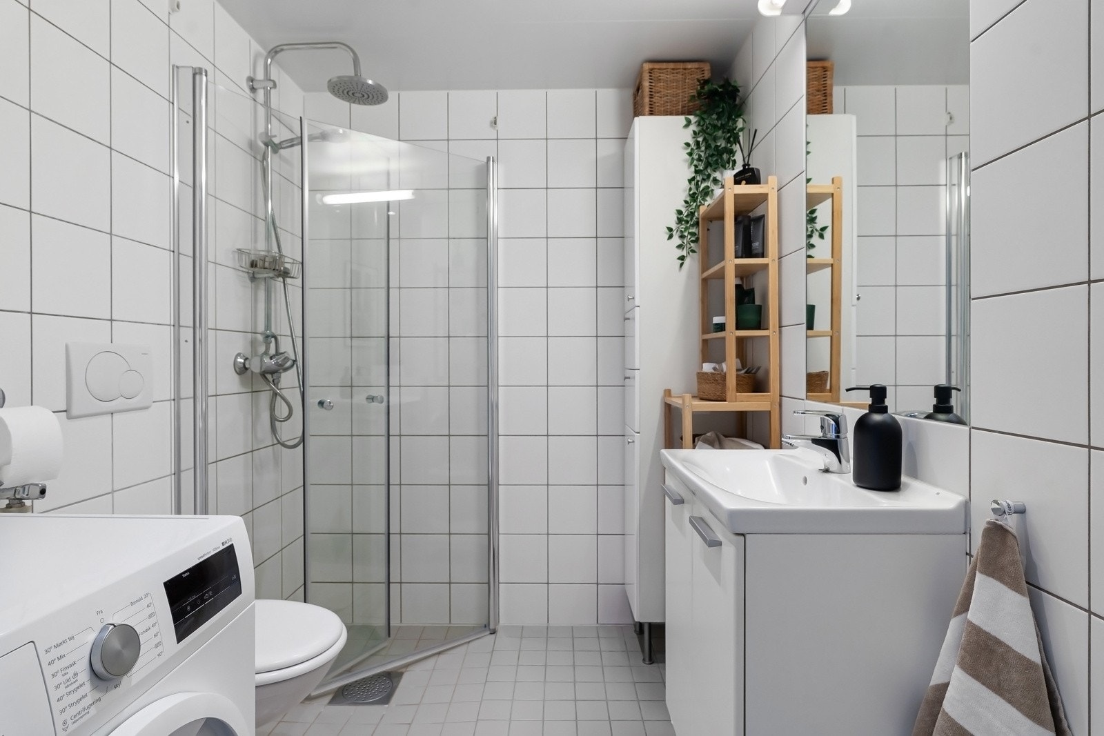 Lyst flislagt bad med nedsenket dusjhjørne med svingbare glassdører, vegghengt toalett og opplegg for vaskemaskin Galleribilde