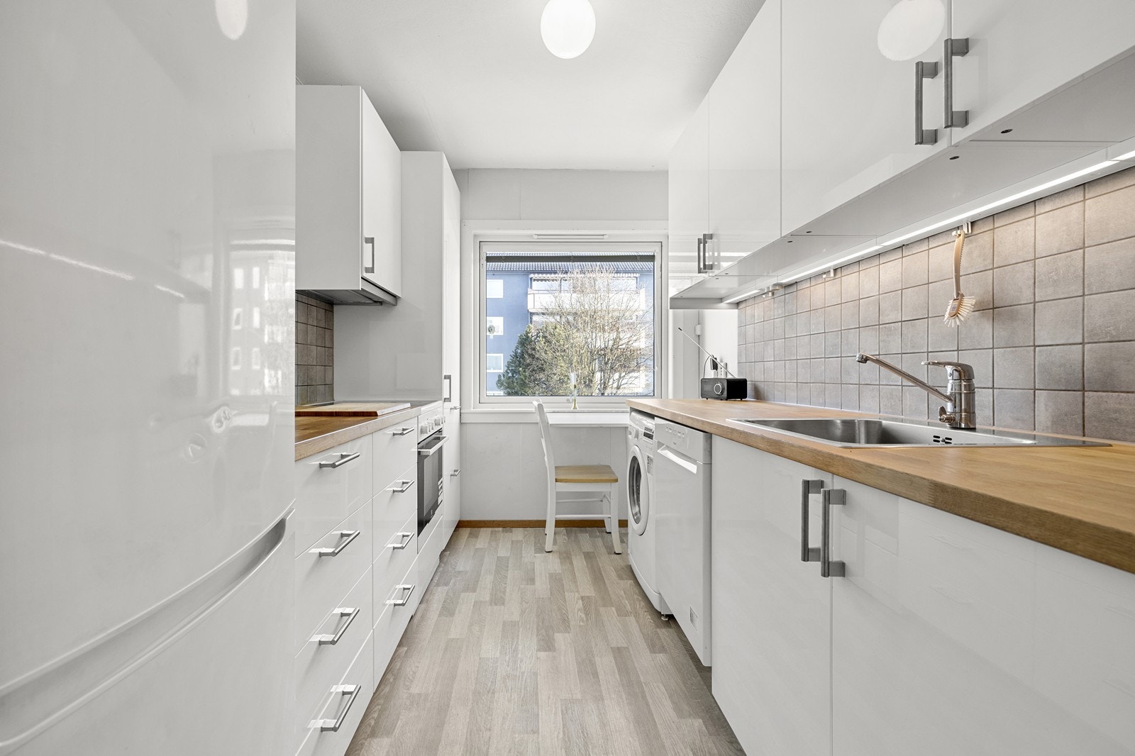 Praktisk kjøkkenløsning som gir mye skap- og benkeplass. Innredningen er hvit med høyglans fronter, det er flislagt sprutsone og belysning under overskap. Galleribilde