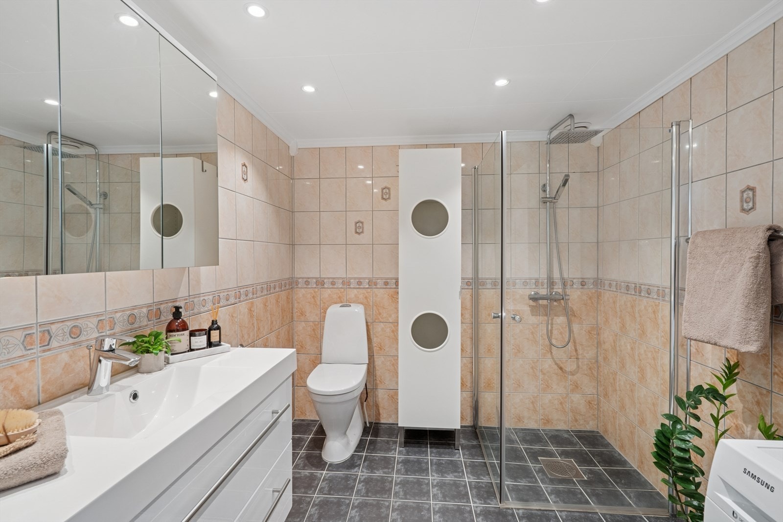 Romslig, flislagt baderom med varmekabler i gulv. Badet har dusjhjørne med nyere glassdører og regnfalls- dusj. Videre er det godt med skapplass i både speilskap og servantinnredning. Opplegg for vaskemaskin og tørketrommel i søyle. Galleribilde