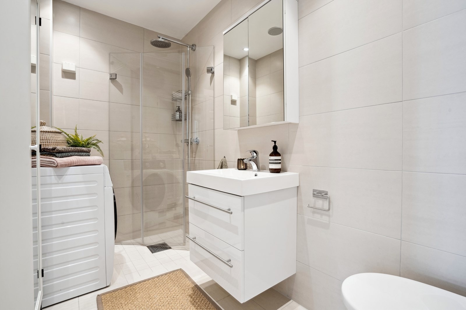 Badet er utstyrt med dusjvegger, vegghengt toalett, servantinnredning med slette overflater, overhengende speil, samt opplegg for vaskemaskin. Galleribilde