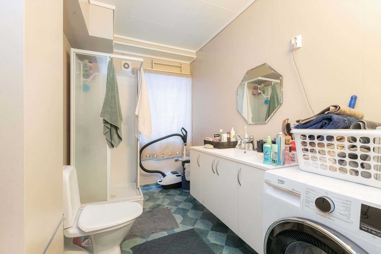 Romslig bad av eldre standard, utstyrt med toalett, dusjkabinett og opplegg for vaskemaskin, samt servantskap med heldekkende servant. Galleribilde