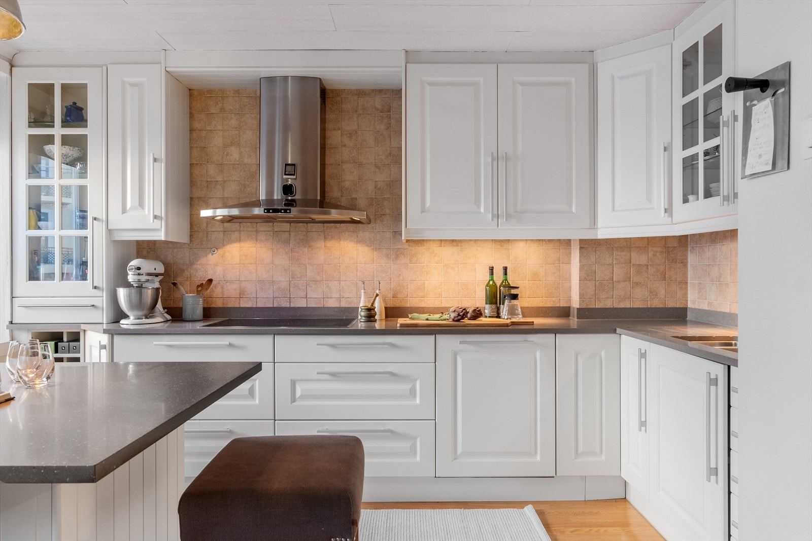 Kjøkkeninnredning med profilerte fronter, benkeplate av stein med nedfelt vask-og skyllekum, samt fliser og belysning over benkeplaten. Galleribilde