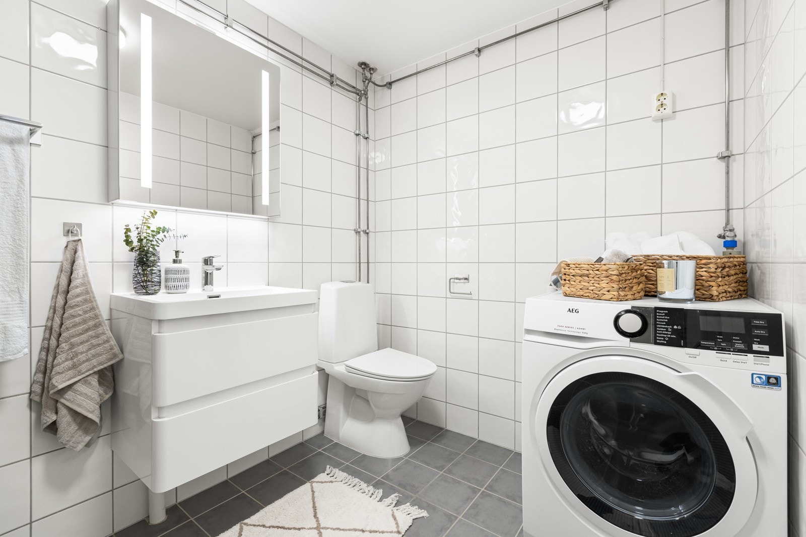 Lyst flislagt baderom med glatt baderomsinnredning med heldekkende servant nedfeldt i underskap, speilskap m/integrert lys, toalett og opplegg for vaskemaskin Galleribilde