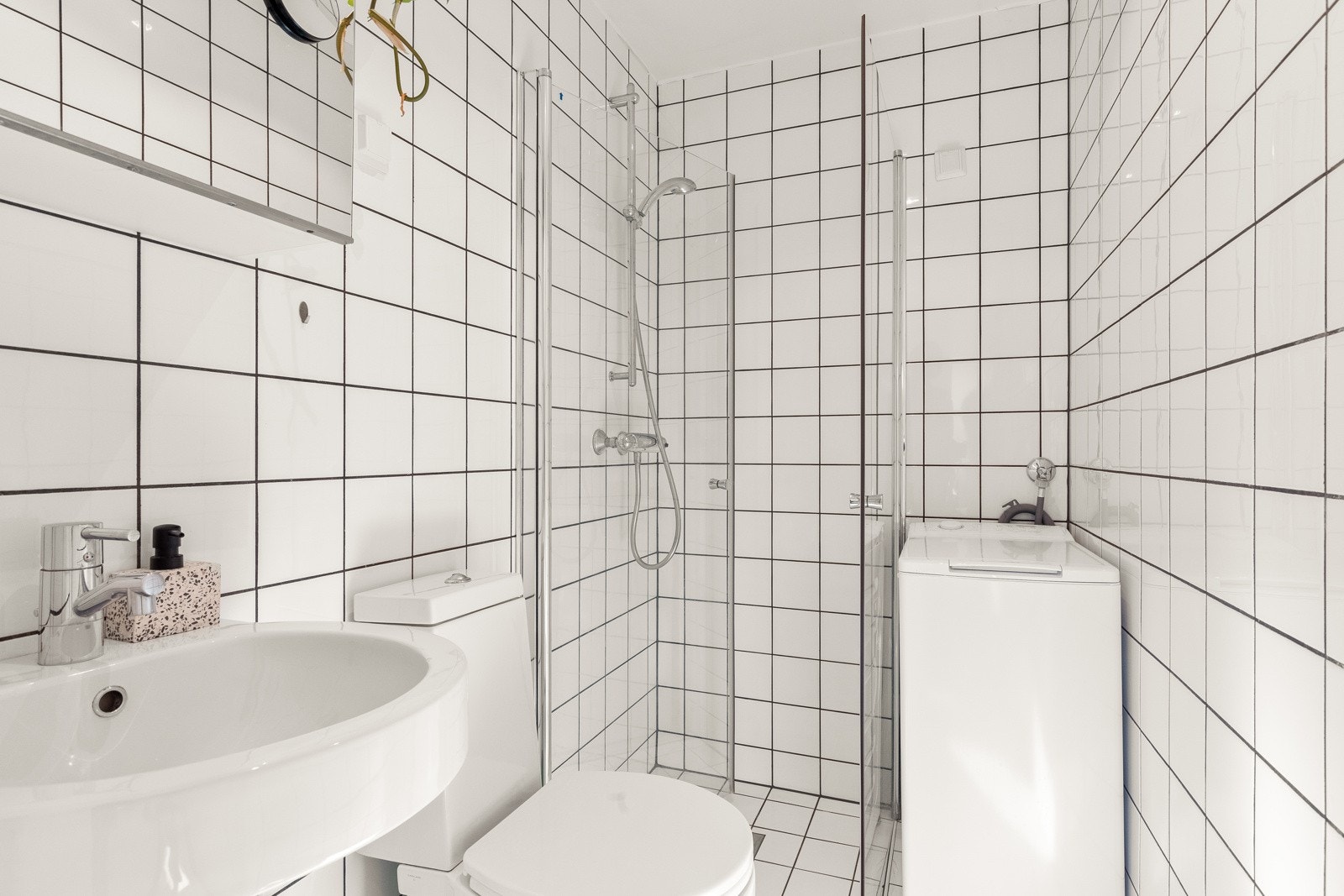 Pent, flislagt baderom som inneholder dusjhjørne, vegghengt servant, toalett og opplegg for vaskemaskin. Galleribilde