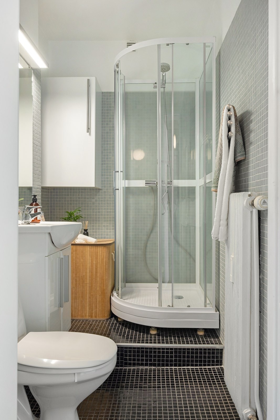 Flislagt baderom med radiator på vegg og mosaikk fliser. Baderommet er innredet med klosett, servant som er nedfelt i innredning, speil og dusjkabinett. Galleribilde