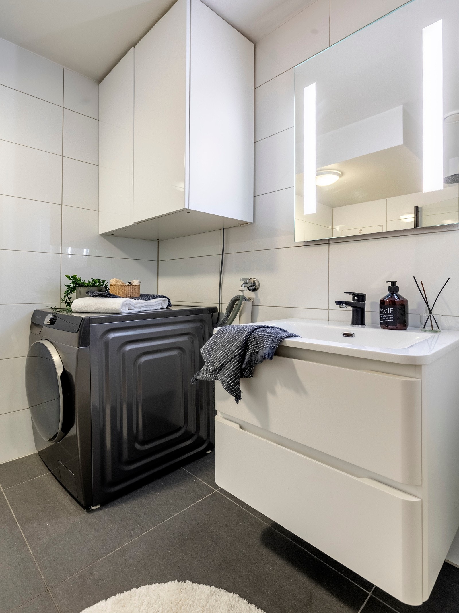 Innredningen inkluderer en servantinnredning, speil med lys på vegg over servant, overskap på vegg over plass for vaskemaskin, vegghengt toalett og dusjhjørne med svingbare glassdører. Galleribilde