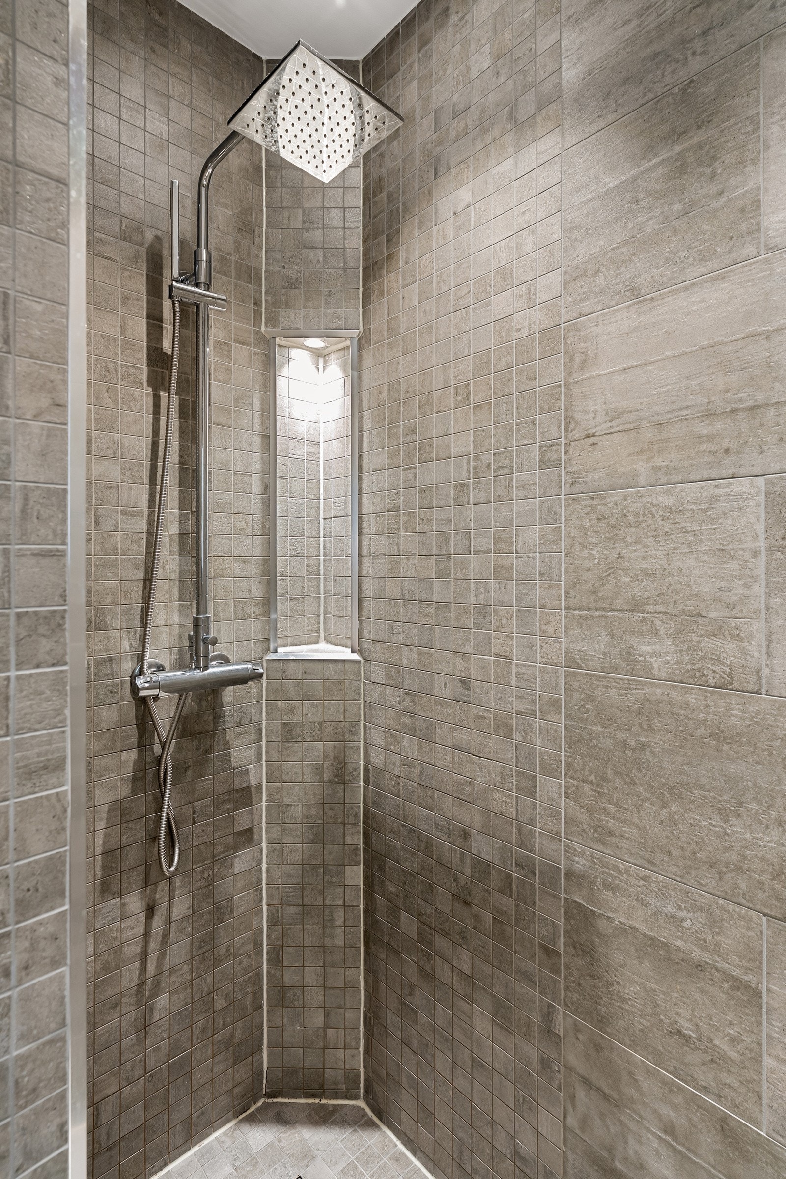 Badet er innredet med en nedsenket dusjsone med regnfallsdusj. De store, pene grå flisene harmonerer med de mindre flisene i dusjsonen, noe som skaper et gjennomført uttrykk. Galleribilde