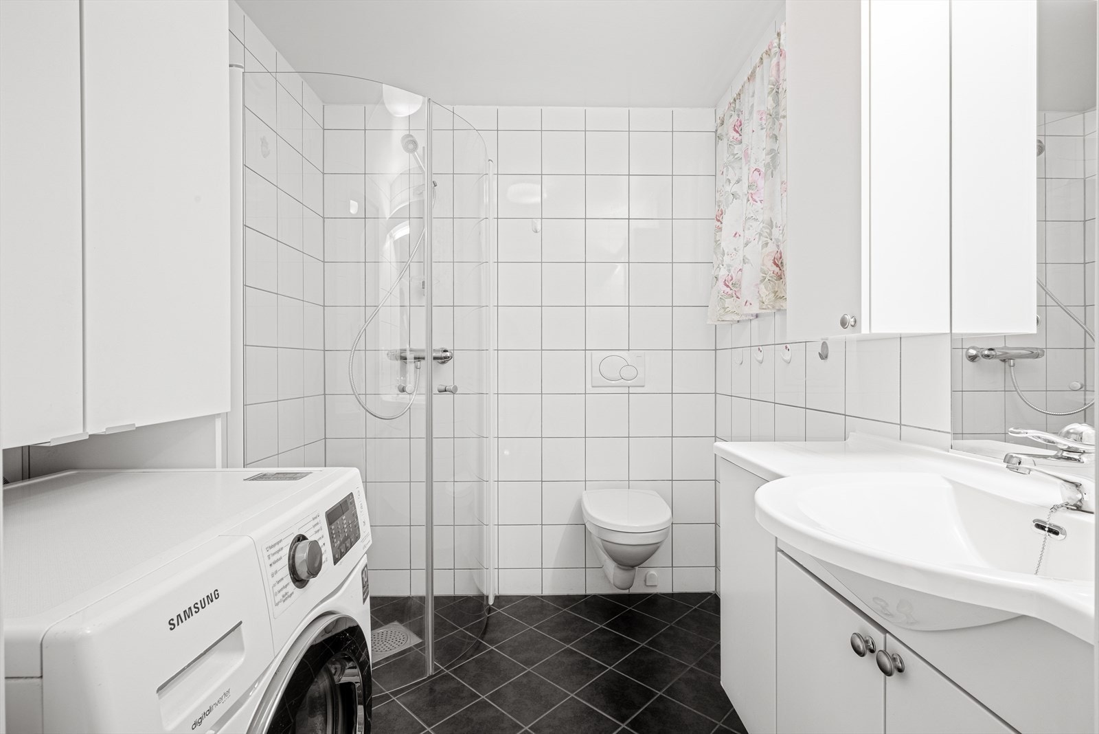 Badet har flislagt gulv med gulvvarme. Lys innredning med over- og underskap, vegghengt toalett, innsvingbare dusjdører og opplegg for vaskemaskin. Galleribilde