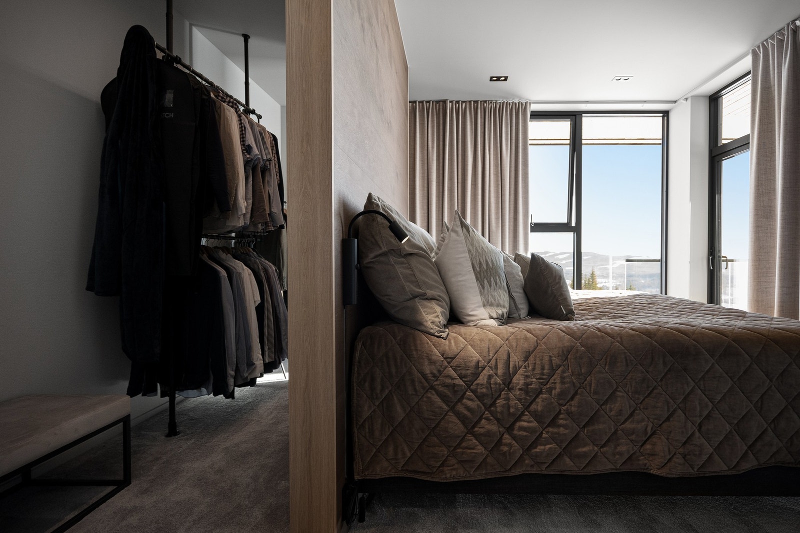 Rommet har lyse vegger, teppebelagt gulv. Det er en åpen garderobeløsning med hengende klær og inngang til eget bad. Galleribilde