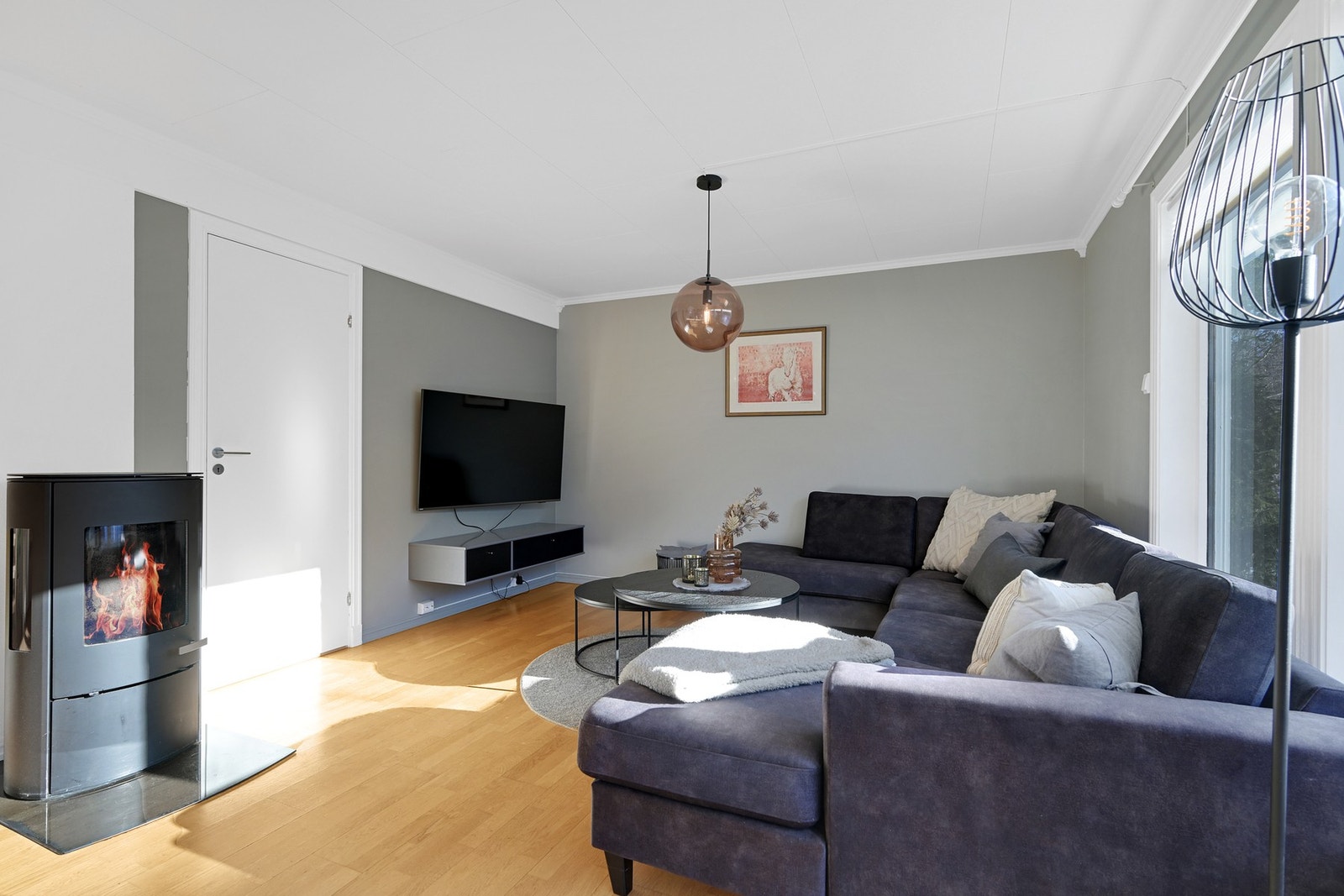 Stuen har lysmalte vegger, store vinduer og parkettgulv. Peisovn i hjørnet gir ekstra varme og hygge. Galleribilde