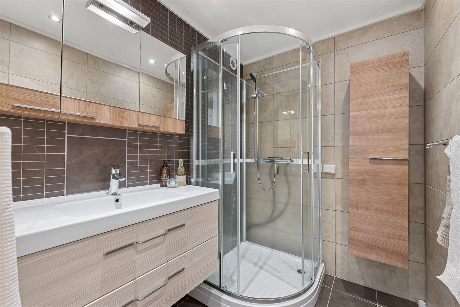 Bad med fliser på vegger og gulv med gulvvarme. Badet har innredning med nedfelt servant med skuffeseksjon, skap og dusjkabinett. Galleribilde