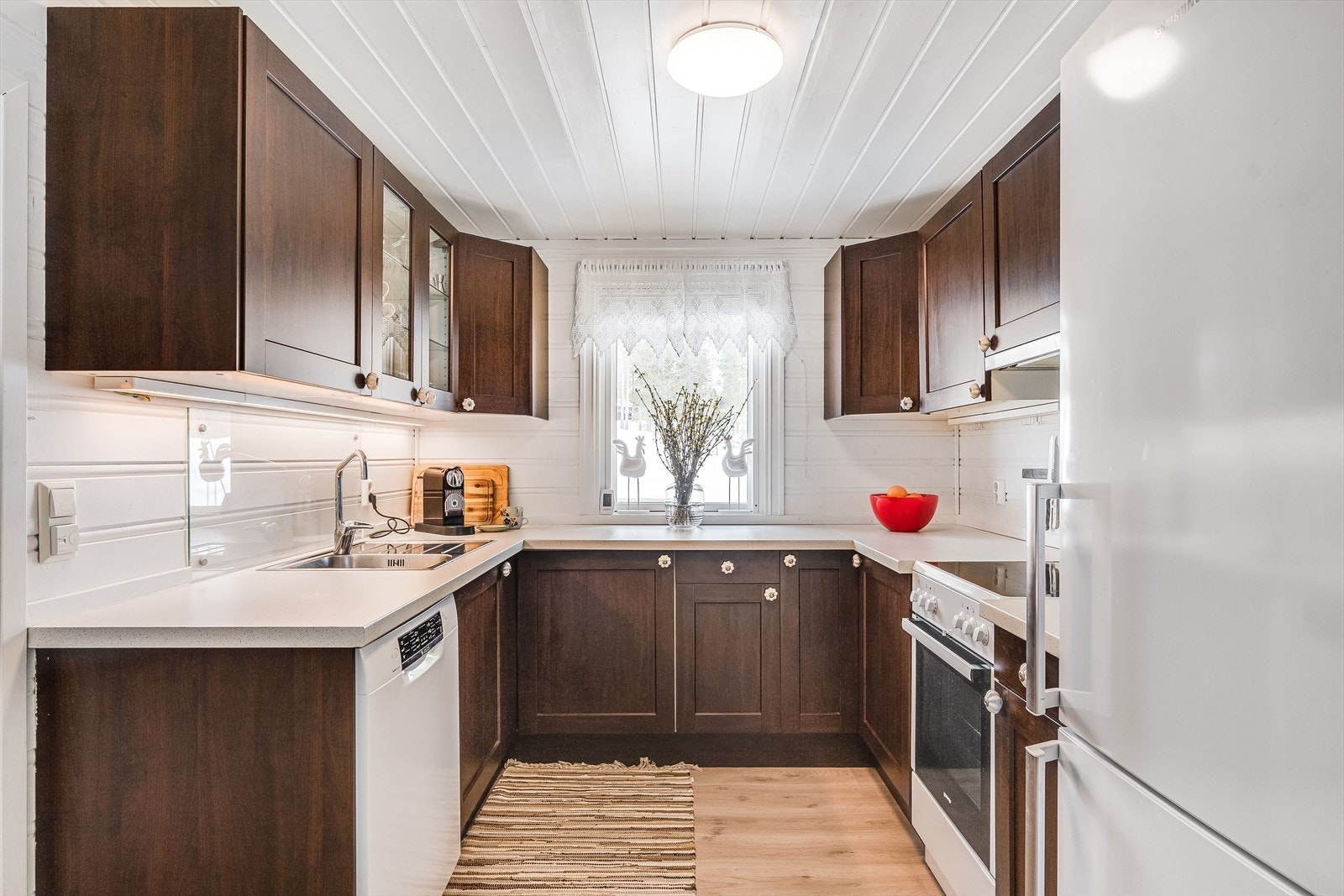 Innenfor gangen finner du et romslig og fint kjøkken som passer godt inn i hyttestilen. Kjøkkenet har rette profilfronter og noen glasskap. Stor laminert benkeplate gir fine arbeidsflater. Merk også vinduet som gir en trivelig arbeidsplass Galleribilde