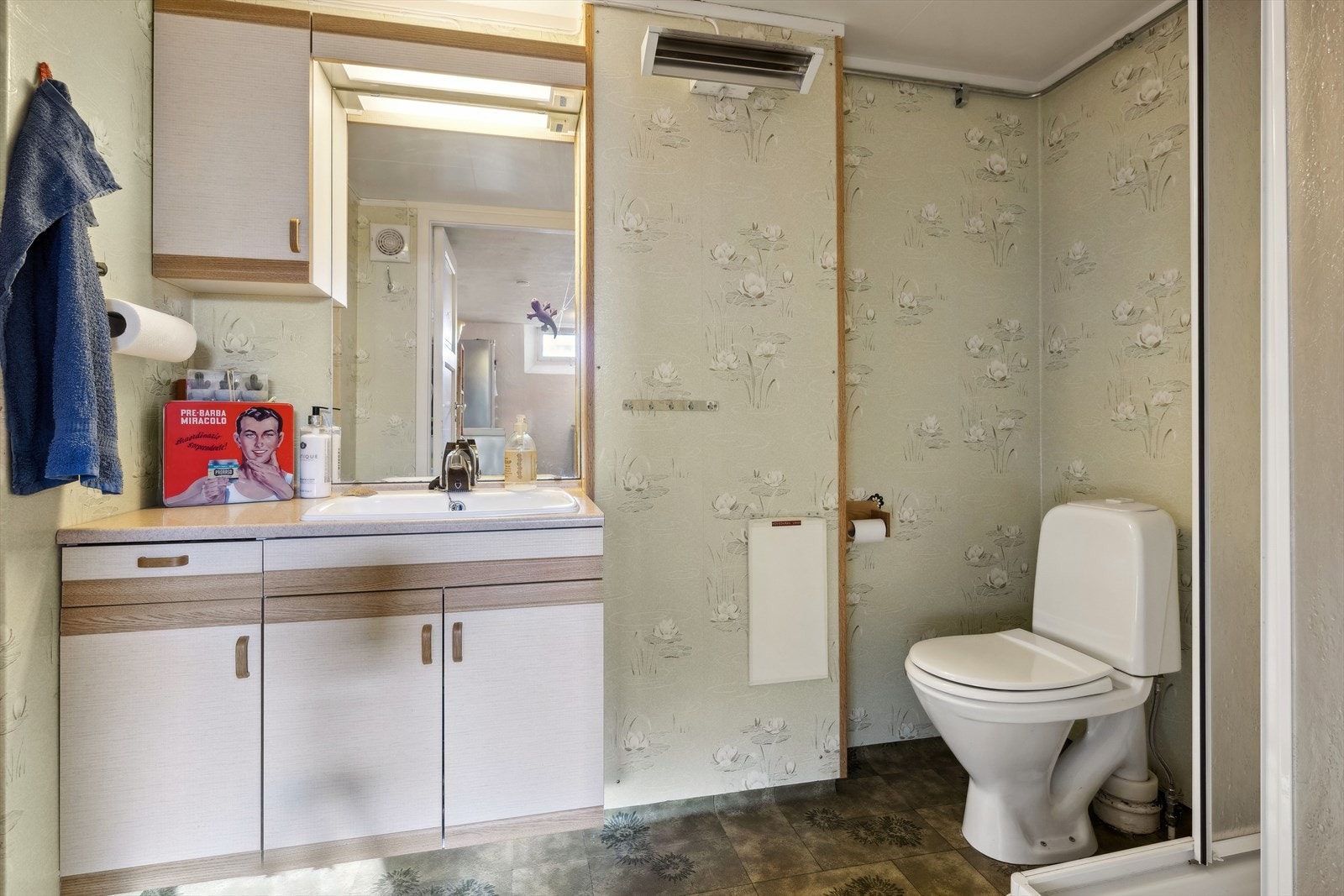 Bad i kjeller med belegg på gulv og vegger med tapet. Badet er utstyrt med toalett, servantinnredning og dusjkabinett. Galleribilde