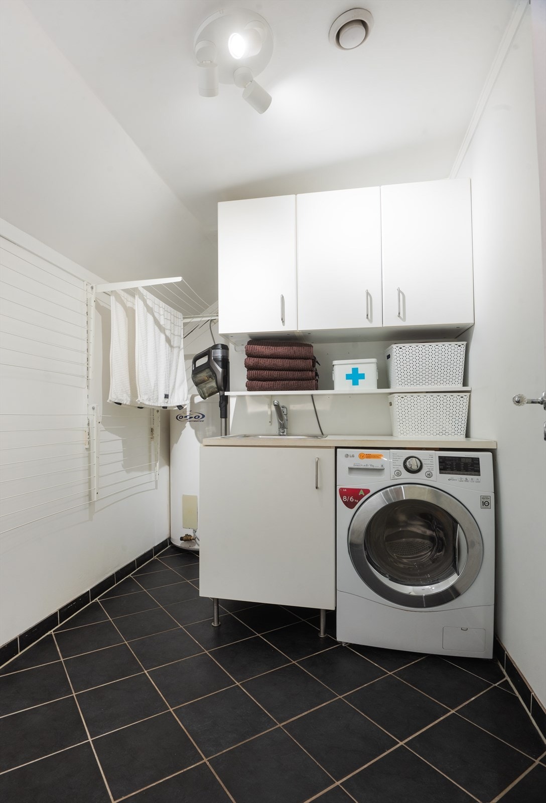 Vaskerom med benkeplate med nedfelt utslagsvask, benkeskap, veggskap og opplegg for vaskemaskin. Galleribilde