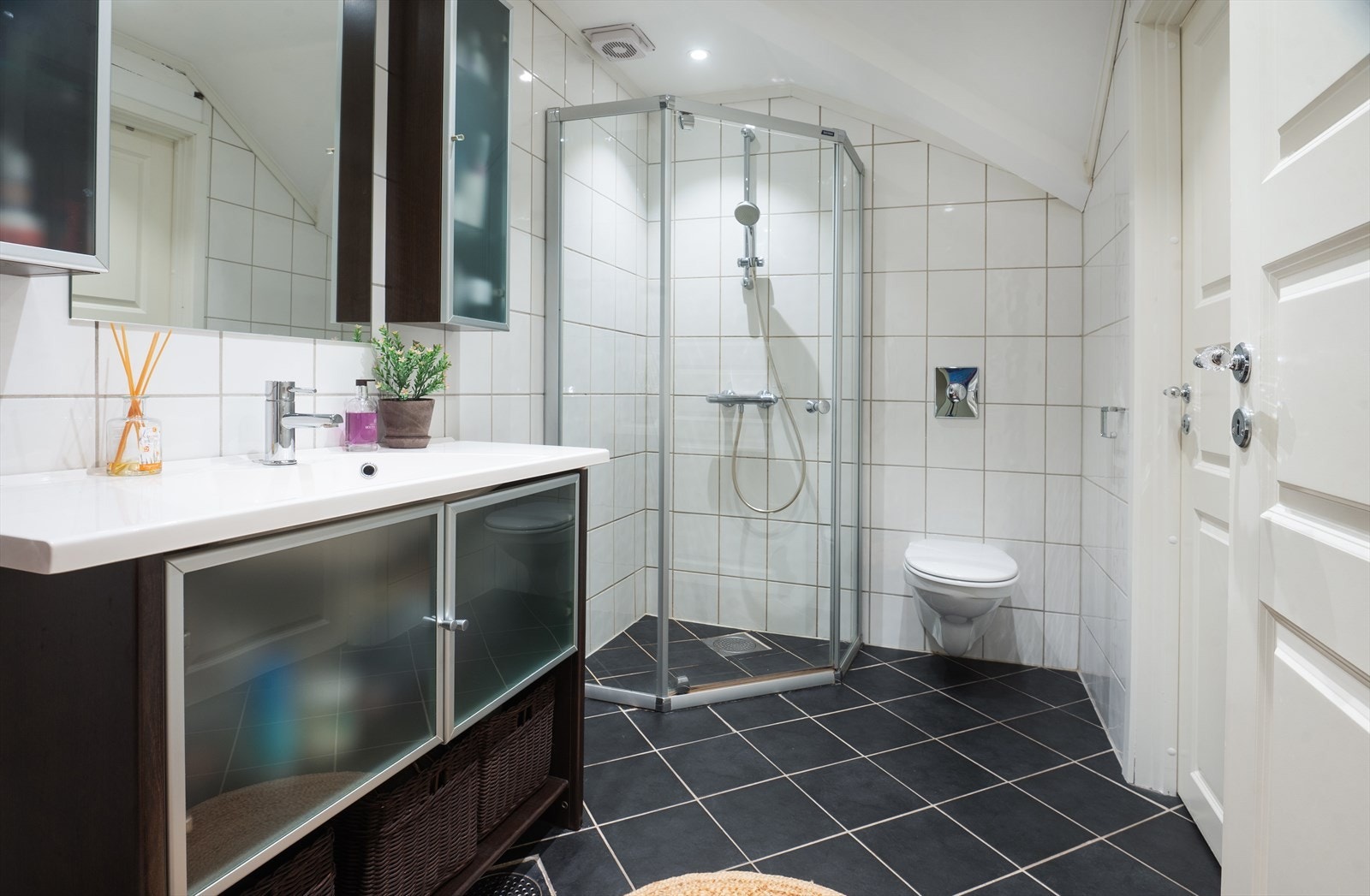 Badet er utstyrt med dusjhjørne med dører i herdet glass, helservant med underskap, speil og veggskap over servant og veggmontert toalett. Galleribilde