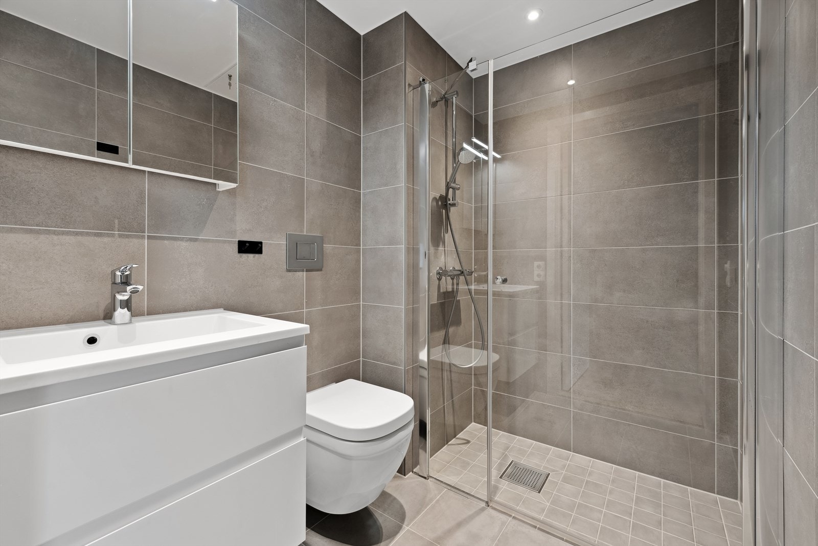 Praktisk løst og pent flislagt bad med lunende varme i gulvet. Dette badet er utstyrt med dusjnisje med glassdør, vegghengt wc og servantinnredning med skuffer og overskap Galleribilde