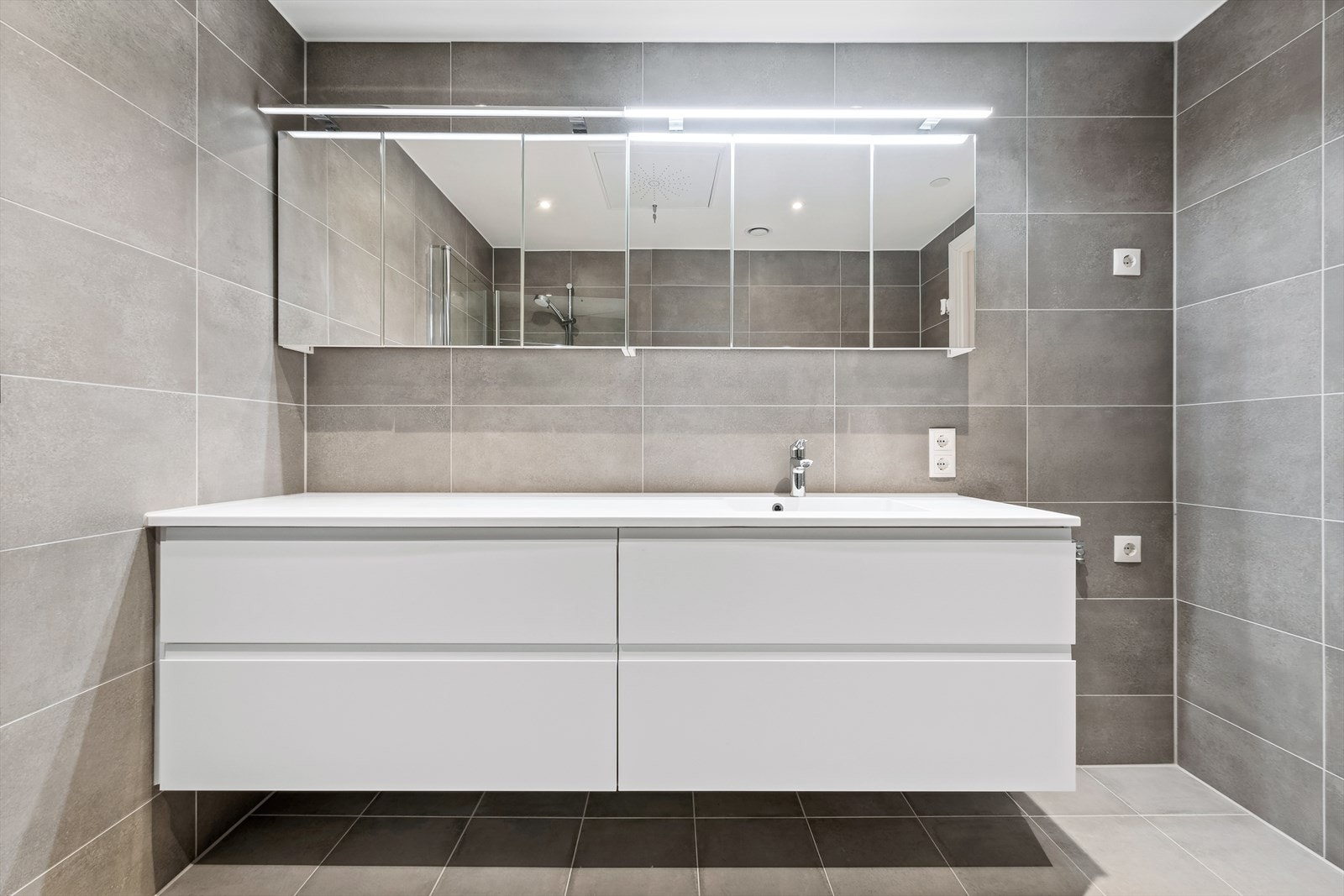 Badet er utstyrt med dusjhjørne med innfellbare glassvegger, vegghengt wc og servantinnredning med skuffer og overskap. I tillegg er det opplegg og plass til både vaskemaskin og tørketrommel Galleribilde