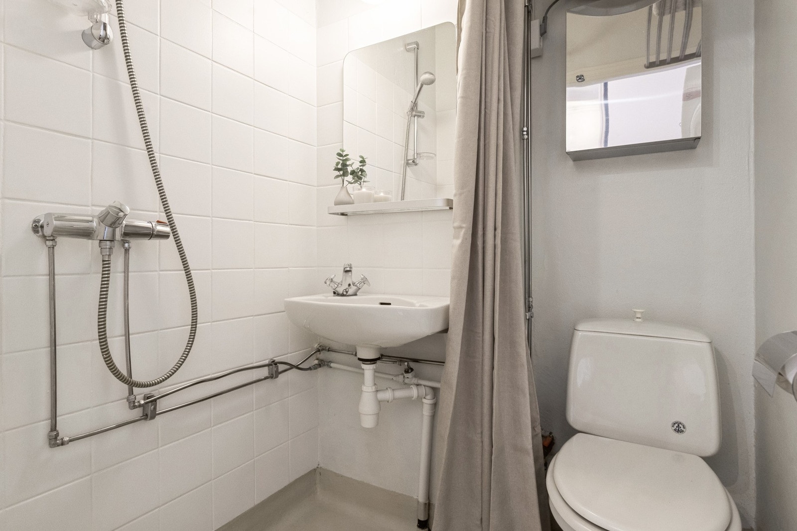 Bad med åpen dusjplass med dusjforheng, dusjgarnityr montert på vegg. Gulvmontert WC. Vegghengt servant. Vegghengt varmtvannsbereder. Galleribilde