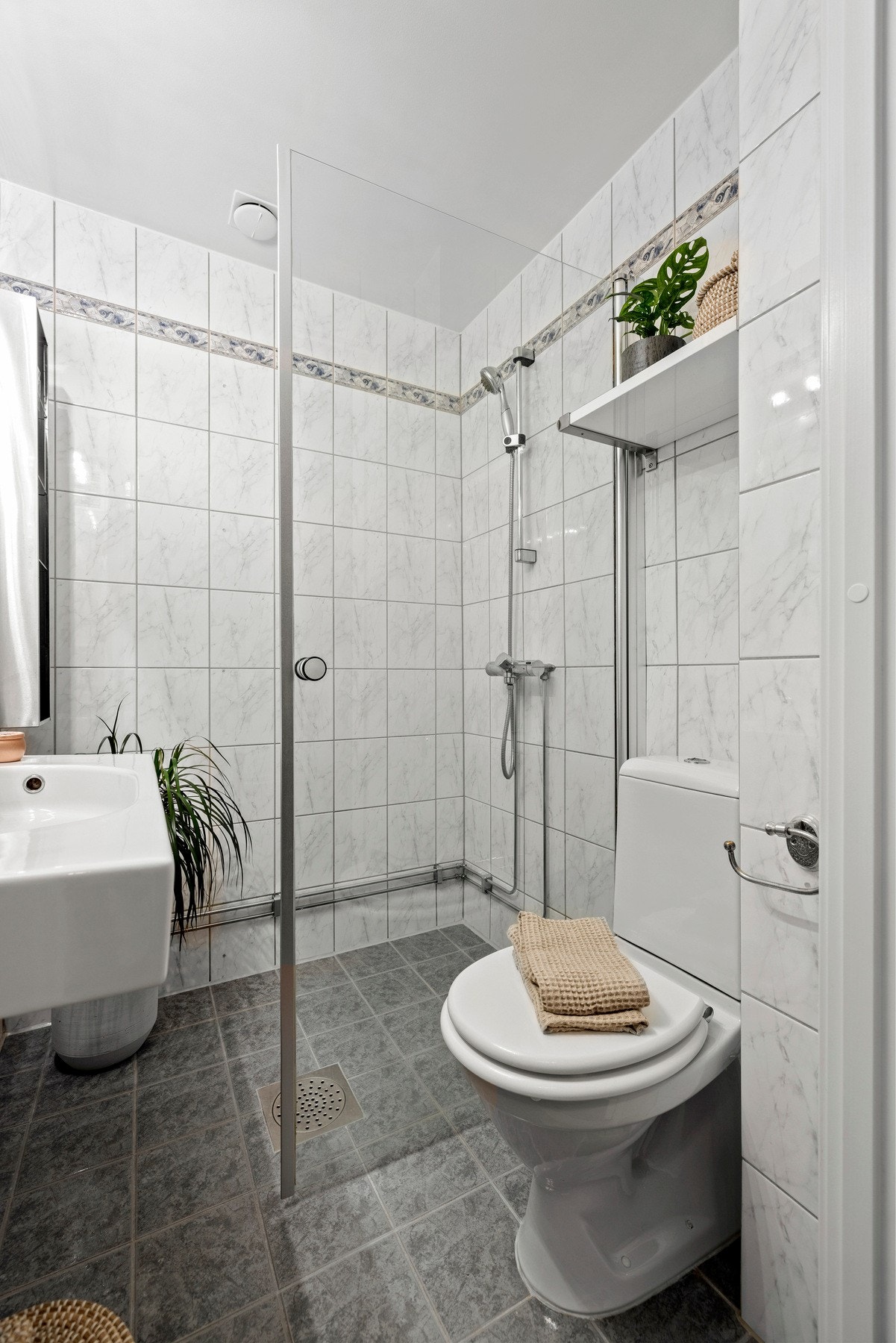 Lyst og flislagt baderom med innredning bestående av vegghengt porselensservant med ettgreps blandebatteri, speil med en lampe montert over, gulvmontert toalett og meget plassbesparende innfellbare dusjvegger. Galleribilde