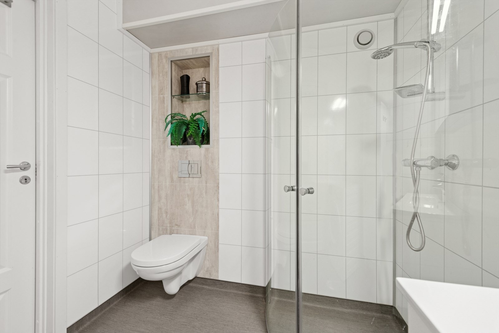 Badet er utstyrt med vegghengt toalett, dusjhjørne med svingbare glassvegg, baderomsinnredning med servant, veggskap, samt opplegg for vaskemaskin og tørketrommel. Galleribilde