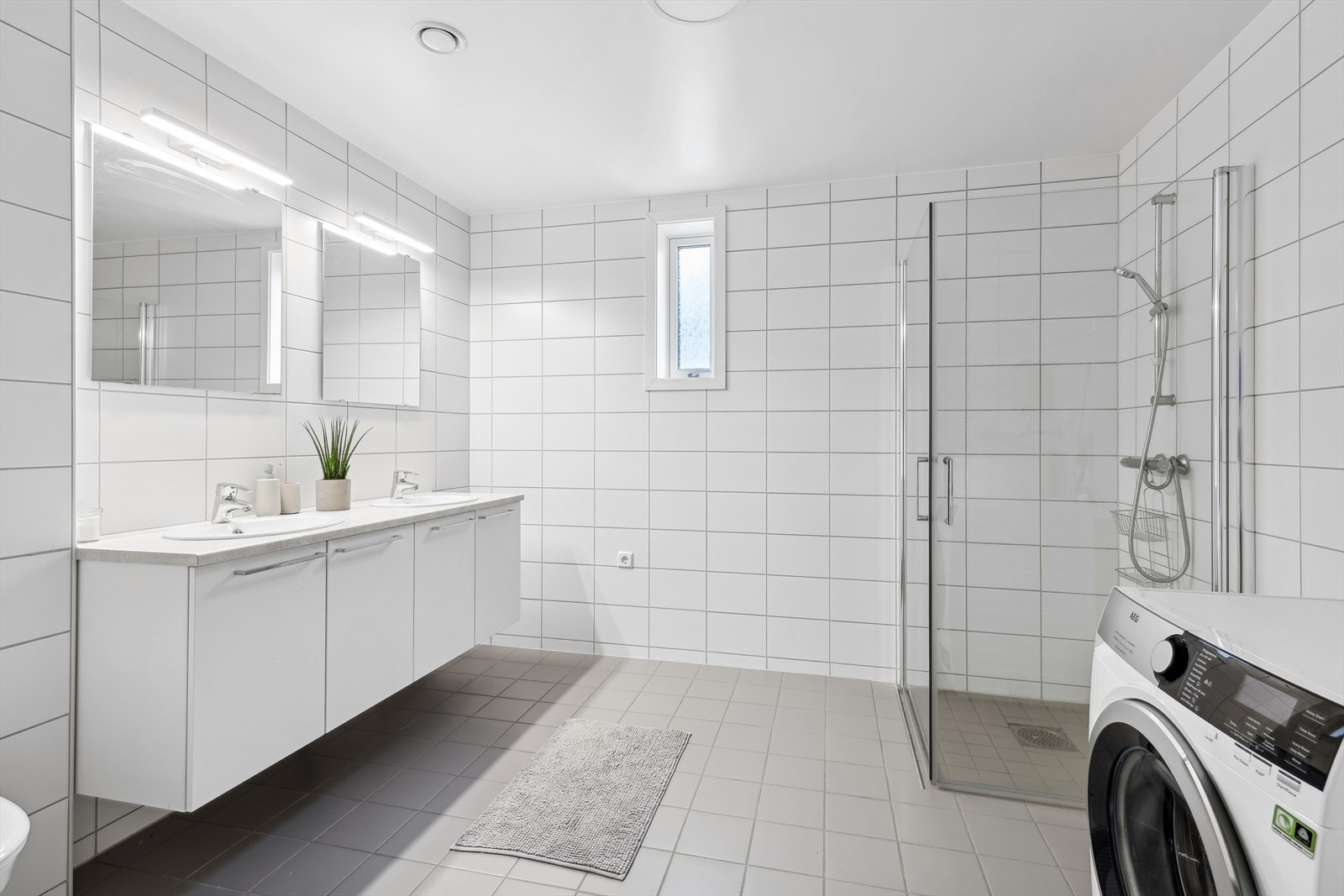 Det flislagte badet har gulvvarme og inneholder et dusjhjørne, vegghengt toalett, samt en innredning med to servanter. Det er også opplegg for vaskemaskin. Galleribilde