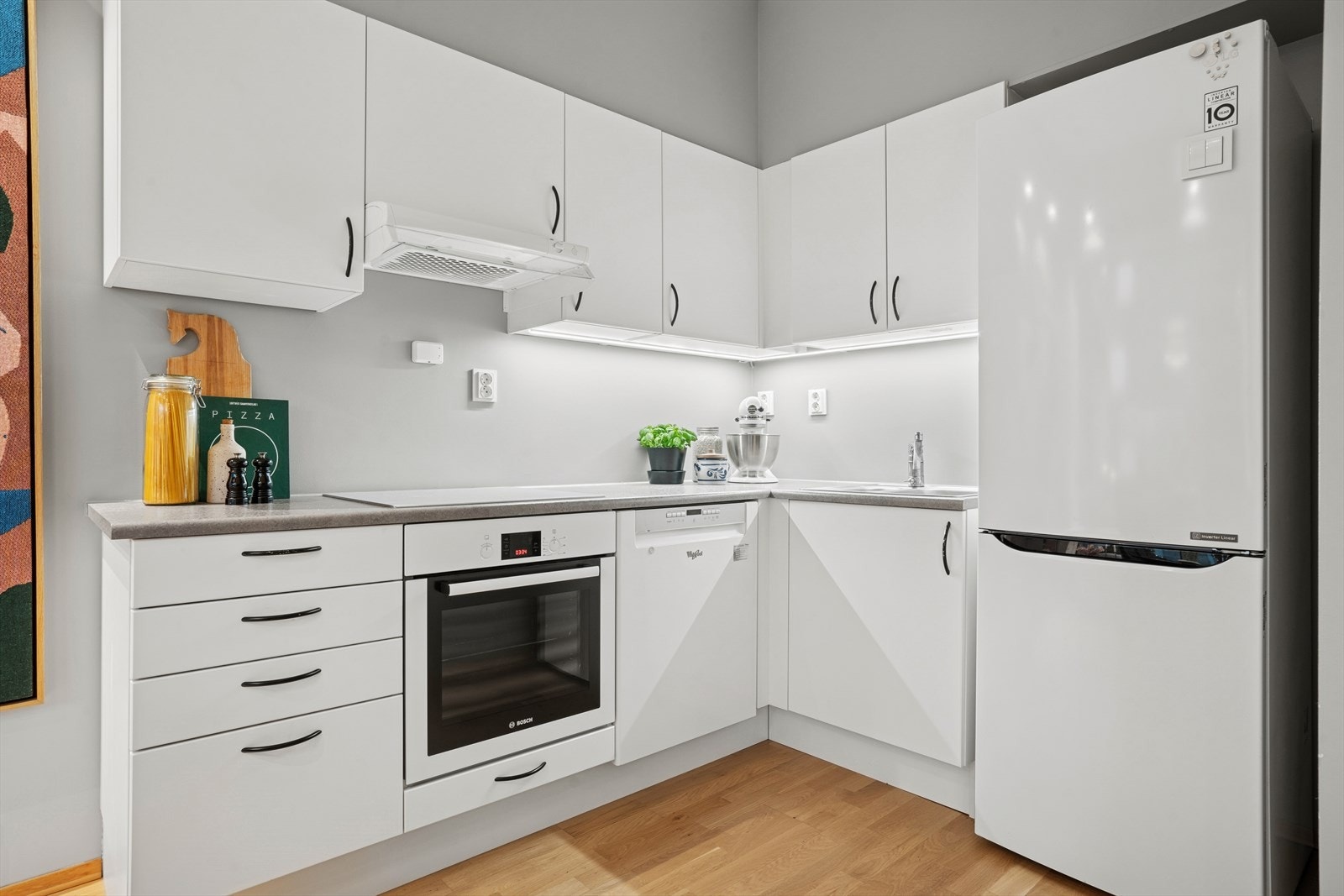 Det tidsmessige Aubo-kjøkkenet er designet i en hendig L-form. Innredningen har hvite, glatte fronter, og grå laminatbenkeplate med nedfelt vaske- og skyllekum. Galleribilde