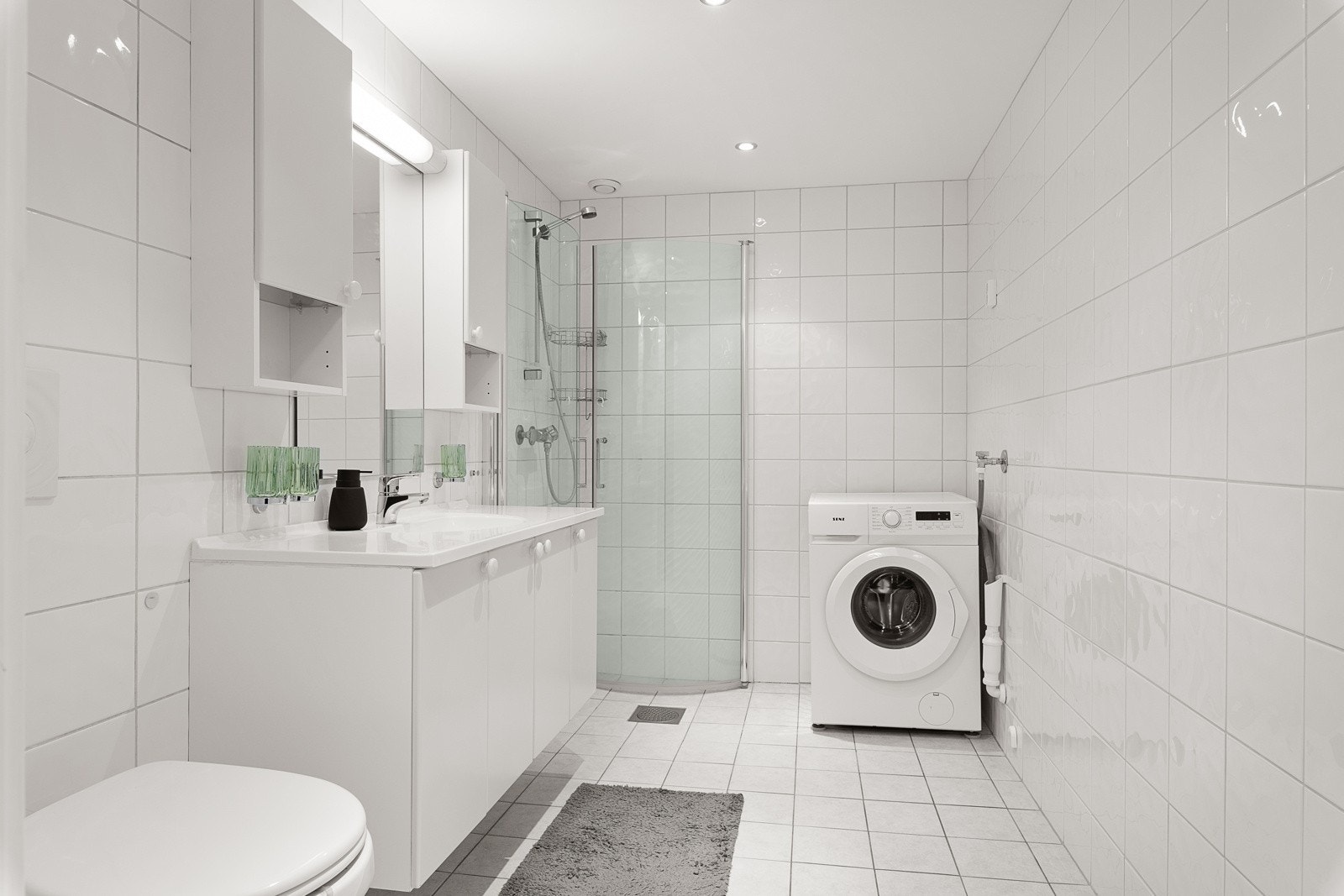 Delikat flislagt bad med dusjhjørne, toalett, servant og opplegg for vaskemaskin Galleribilde