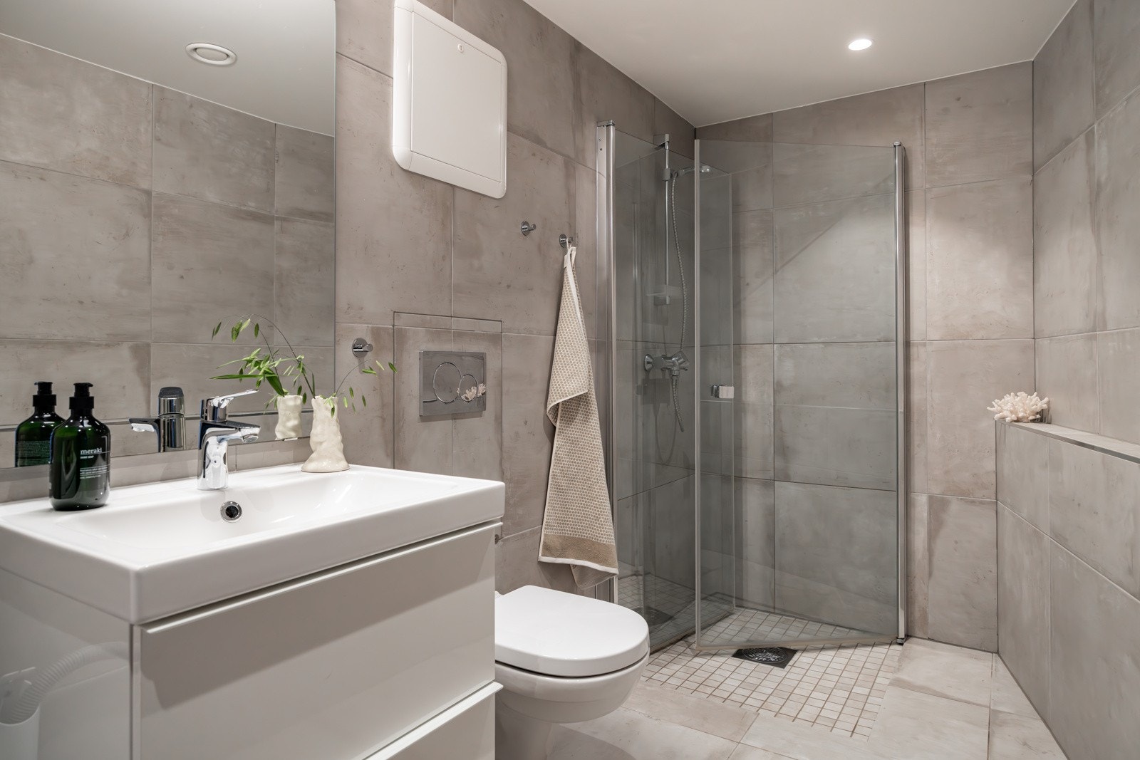 Moderne baderomsinnredning med skuffer og heldekkende servant, speil med belysning over. Baderommet er også utstyrt med dusjhjørne med innfellbare glassdører og vegghengt toalett. Galleribilde