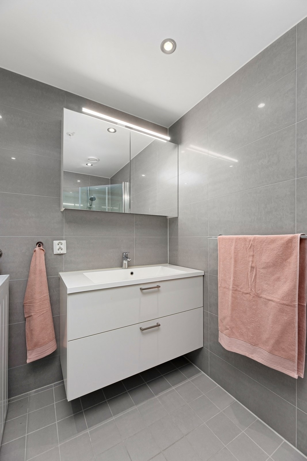 Badet er utstyrt med dusjhjørne med innfellbare glassdører, veggmontert wc, servantskap med hvite laminerte skuffefronter og overmontert hvit servant med ettgreps blandebatteri i rustfritt stål fra Oras. Galleribilde
