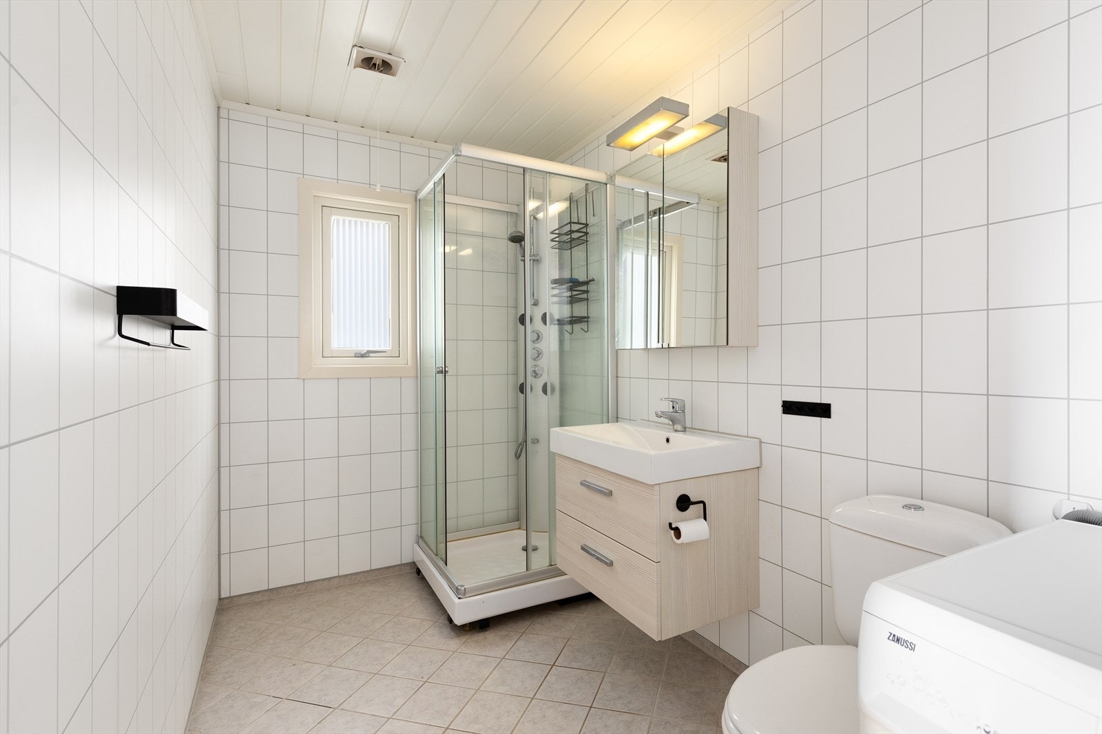 Baderom med fliser på både veggar og golv. badet er utstyrt med dusjkabinett, baderomsinnredning med nedfelt servant, toalett og opplegg for vaskemaskin. Galleribilde