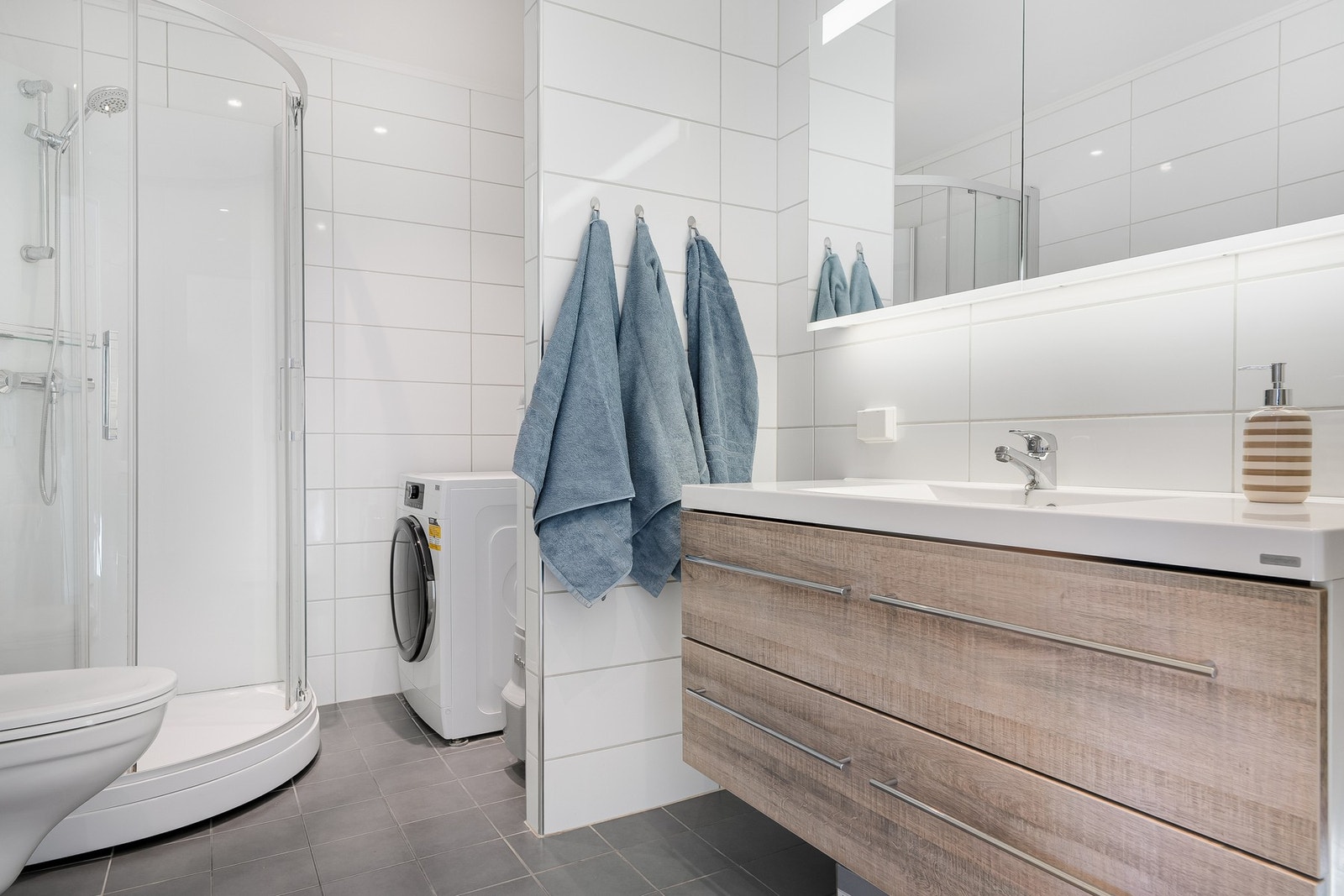 Badet inneholder servant med underskap, vegghengt speil med overlys, toalett og opplegg for vaskemaskin og tørketrommel. Galleribilde
