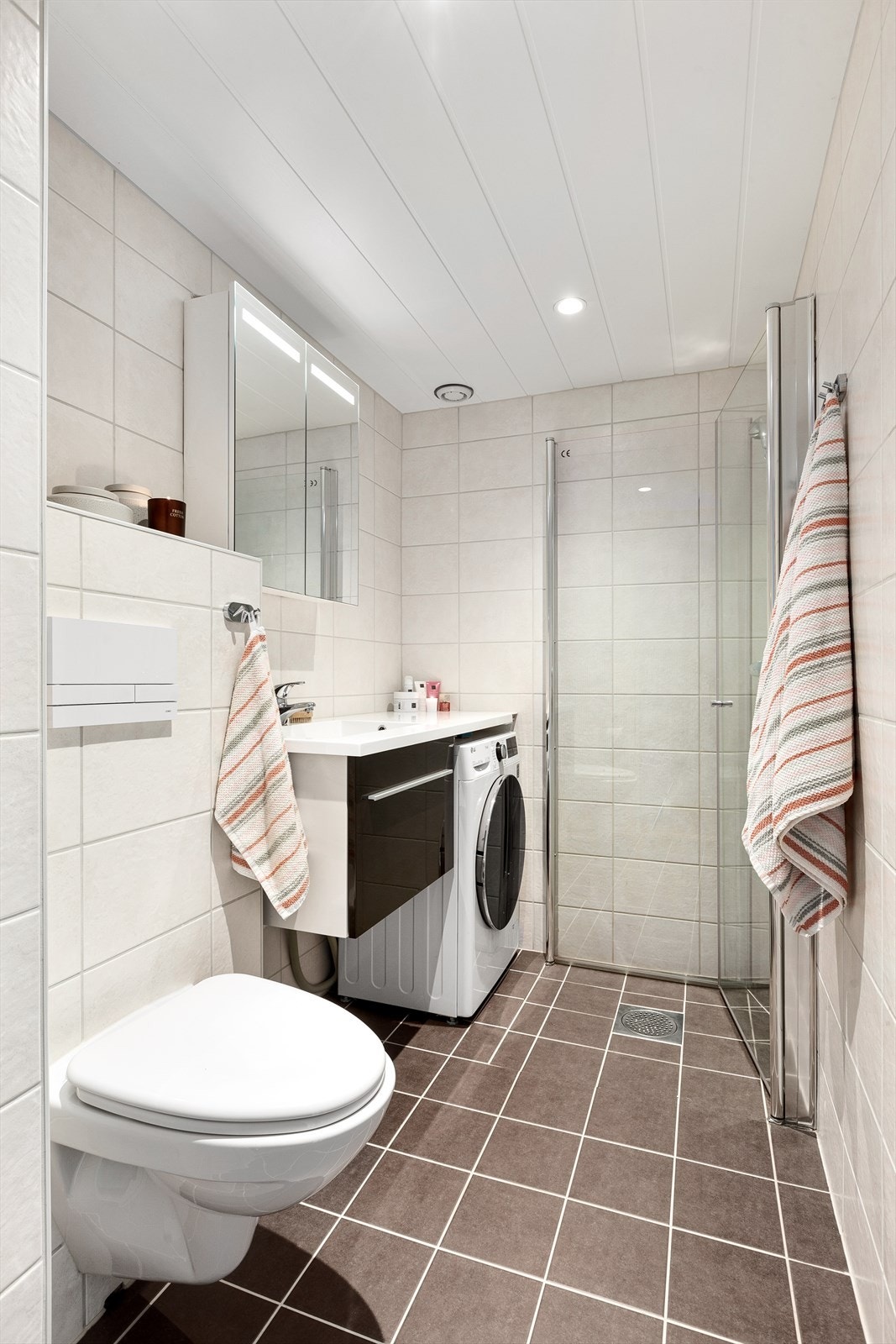 Pent flislagt bad med innredning bestående av klosett, dusjhjørne med innfellbare dører og baderomsinnredning med speilskap og integrerte ledlys Galleribilde