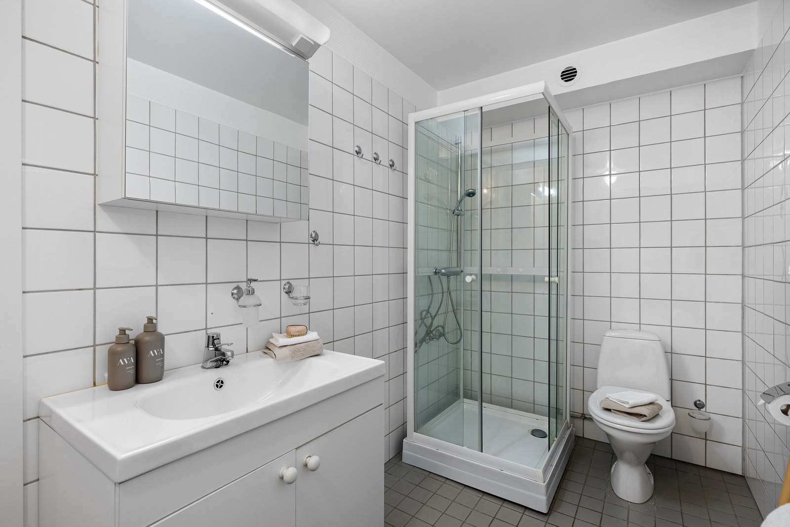 Lyst og flislagt baderom utstyrt med servant, vegghengt speilskap, gulvstående toalett og dusjkabinett. Badet er antatt fra byggeår, og det er iht. tilstandsrapporten gitt TG 3 på rommet Galleribilde