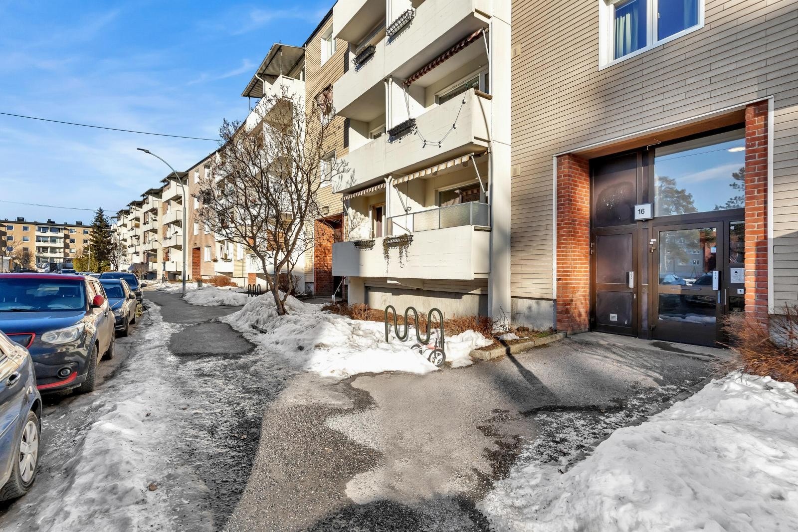 I Bygningen har en lys fasade med teglsteinsdetaljer rundt inngangen. Området er asfaltert og det er parkeringsplasser langs veien. Galleribilde