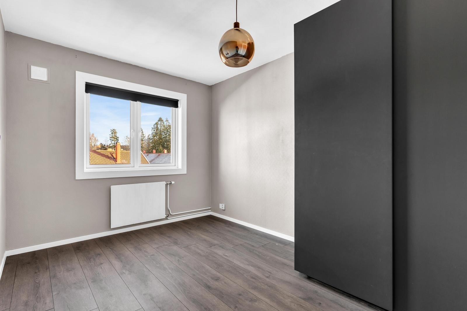 Rommet har grå vegger, mørkt gulv og et stort vindu som slipper inn naturlig lys. Radiator under vinduet. Taklampe i midten av rommet. Galleribilde