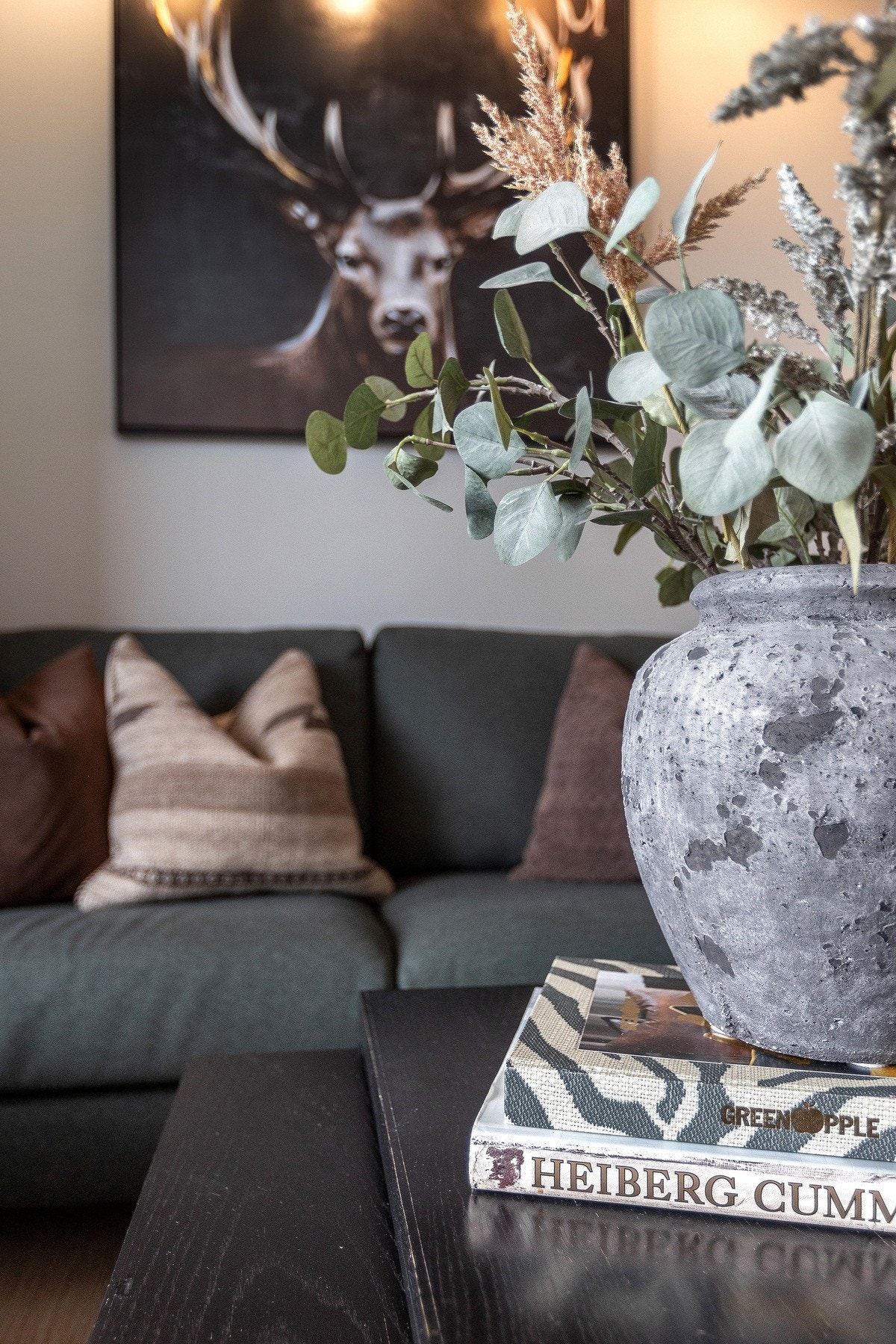 Stuen har en behagelig atmosfære med en mørk sofa og dekorative puter. Veggen har et kunstverk og på bordet står en rustikk vase med tørkede blomster og blader. Galleribilde