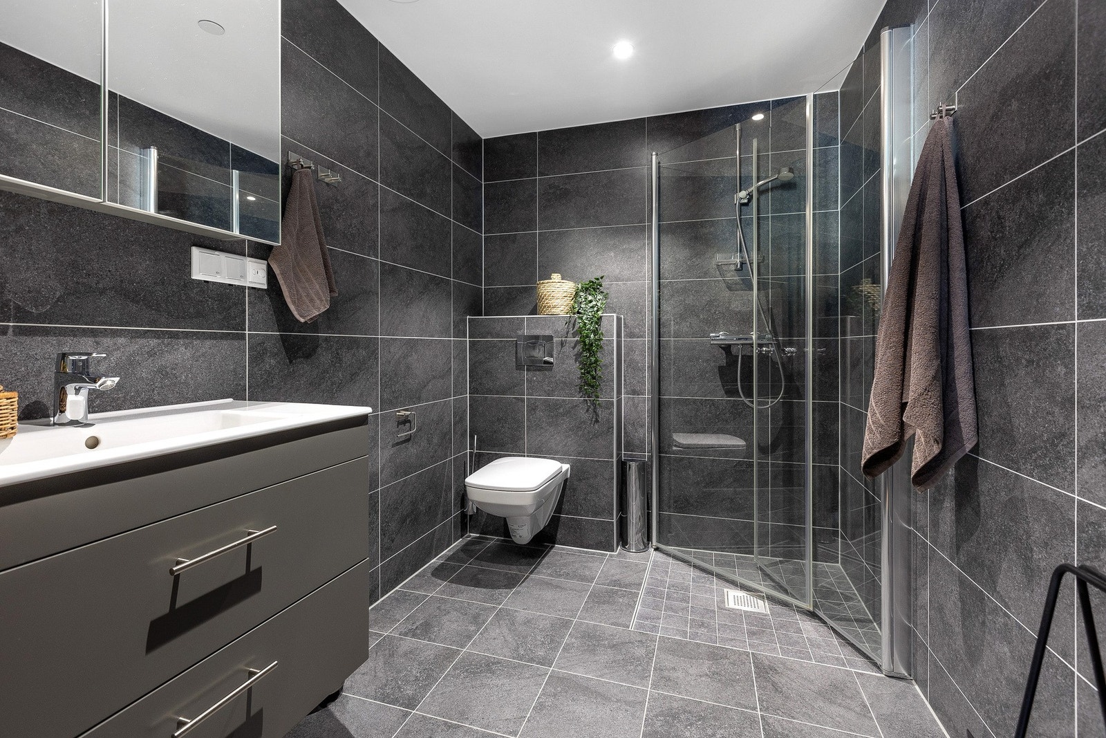 Moderne baderom med mørke fliser på vegger og gulv. Rommet har vegghengt toalett, dusjhjørne med glassdører og en romslig servant med underskap. Downlightsbelysning i taket gir god lyssetting. Galleribilde