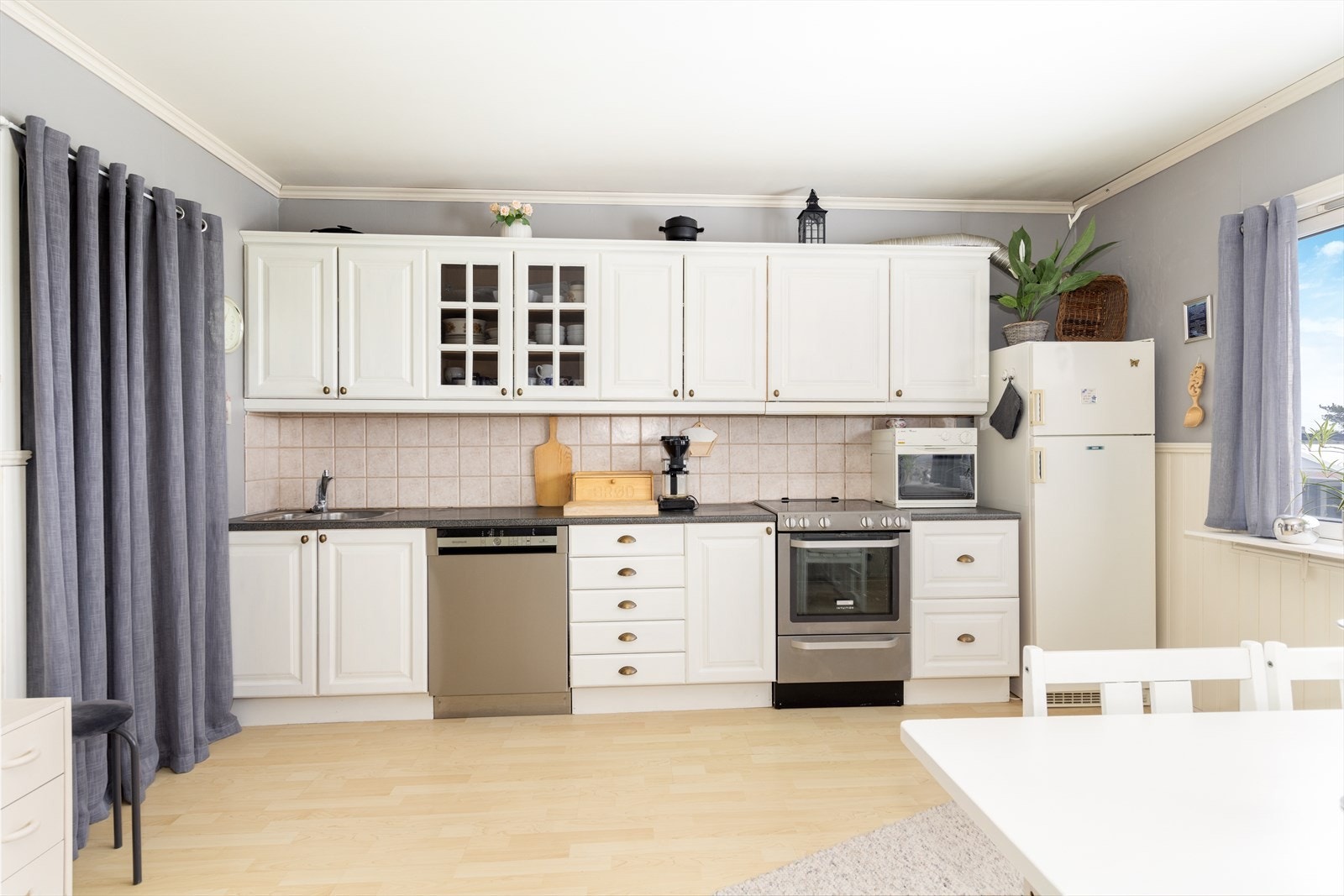 Kjøkkenet har hvit innredning langs den ene veggen fra 2001 med profilerte fronter og innslag av glass-skap. Det er avsatt plass til frittstående hvitevarer som oppvaskmaskin, komfyr og kjøl/fryseskap, og over kokesonen er det ventilator med avtrekk ut. Galleribilde