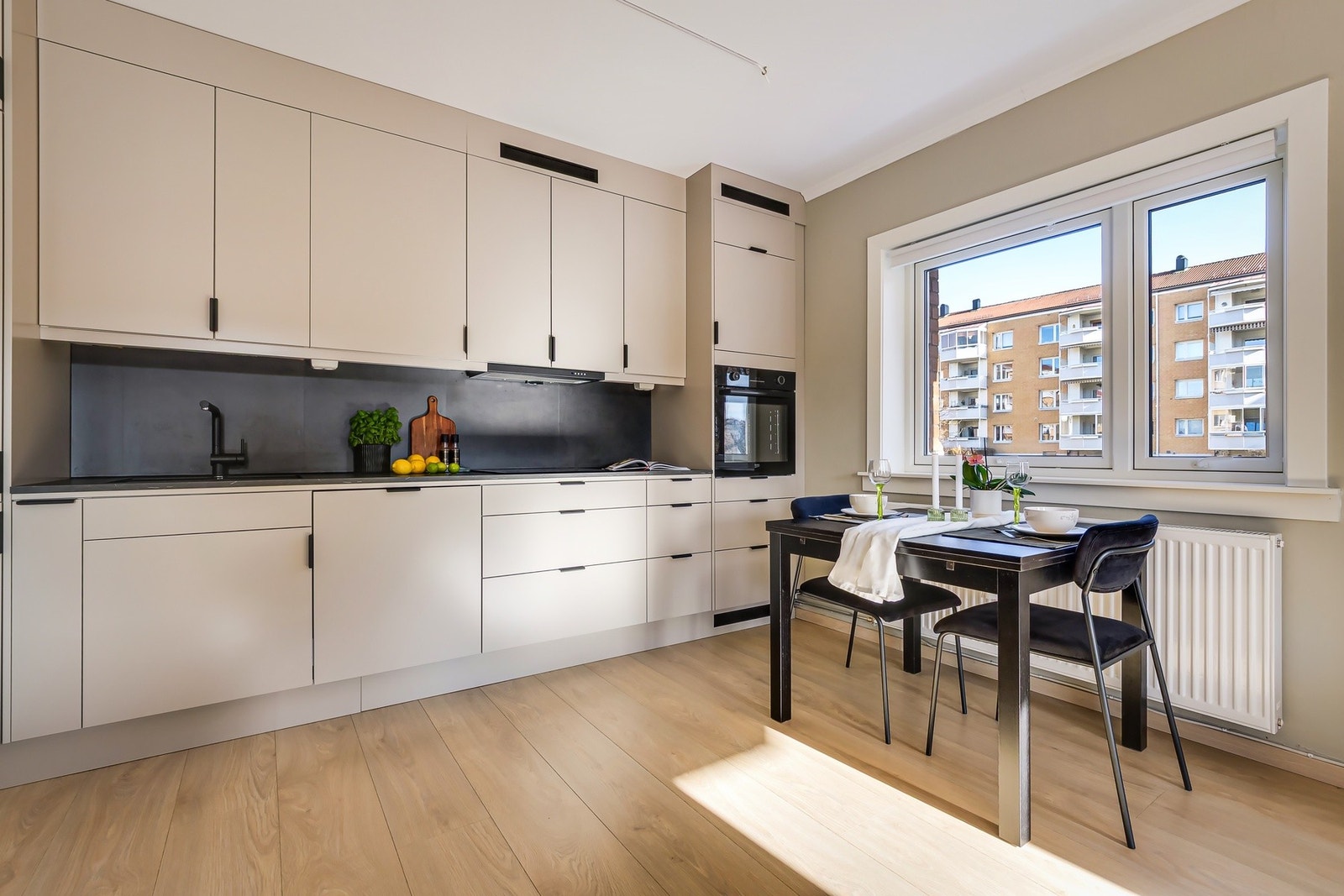 Innredningen har glatte fronter i en lys beige farge, en laminat benkeplate med nedfelt kum i kompositt, sort blandebatteri, belysning under overskap og kjøkkenplater på veggen over benkeskapene. Galleribilde