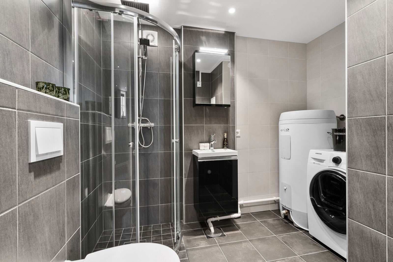 Badet i underetasjen er helfliset med grå fliser og innredet med en tidløs sort innredning, dusjnisje, vaskemaskin og vegghengt toalett. Galleribilde