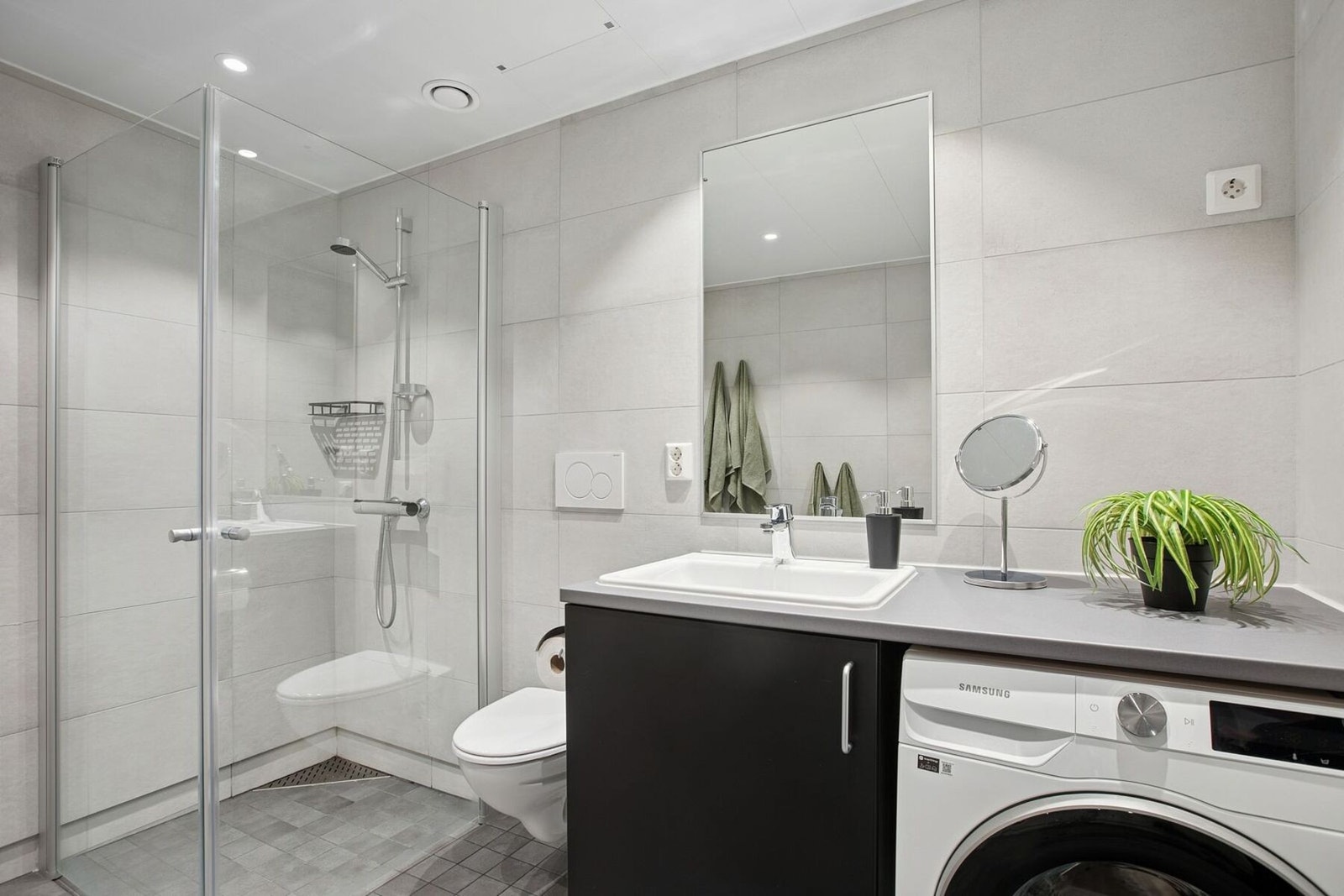 Badet er innredet med servant med underskap og speil, dusjhjørne med innfellbare glassdører i herdet glass, samt vegghengt toalett. Galleribilde