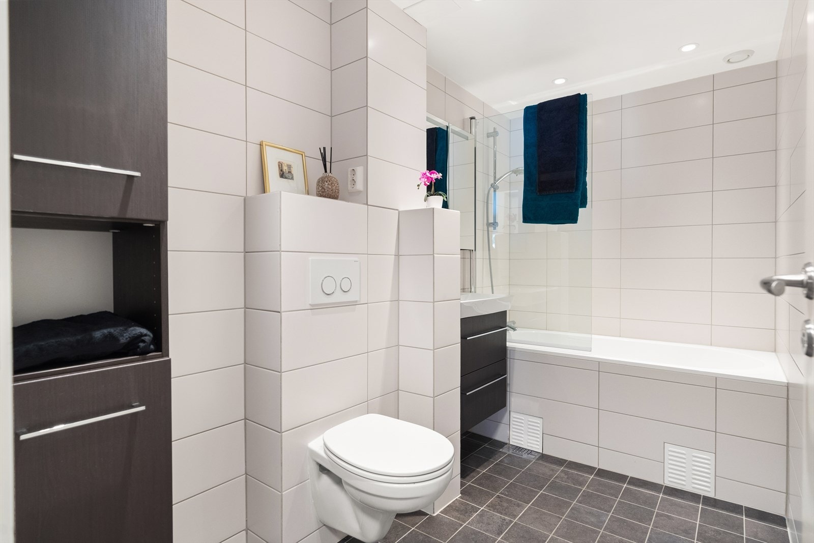 Delikat og tidløst baderom med vegghengt toalett, vegghengt servantinnredning, høyskap og speilskap, samt badekar. Galleribilde