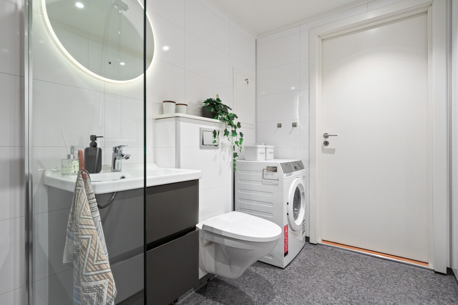 Badet har et moderne baderomsmøbel med grå skuffefronter, et rundt speil med integrert belysning over servanten, veggmontert toalett, og et romslig dusjhjørne med glassvegg og regnfallsdusj. Galleribilde