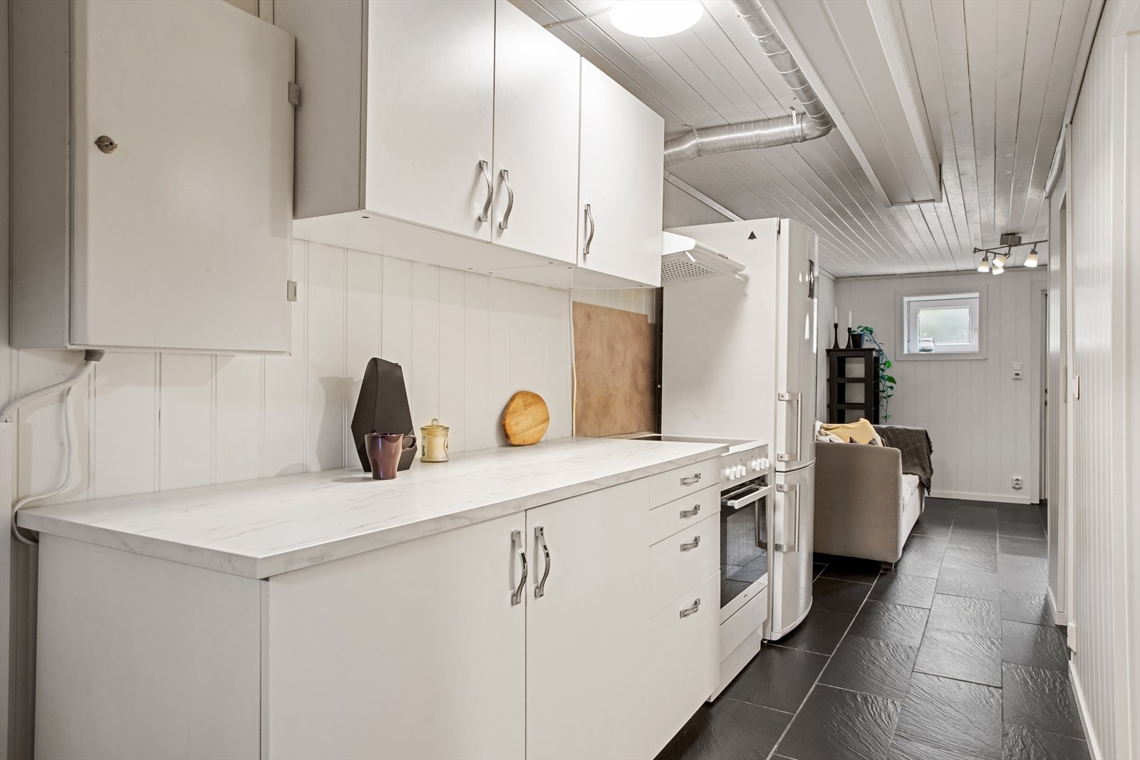 Åpen stue-/ kjøkkenløsning med eldre og noe nyere kjøkkeninnredning. Kjøkkenet har slette og profilerte fronter, benkeplater av laminat og vask av stål Galleribilde