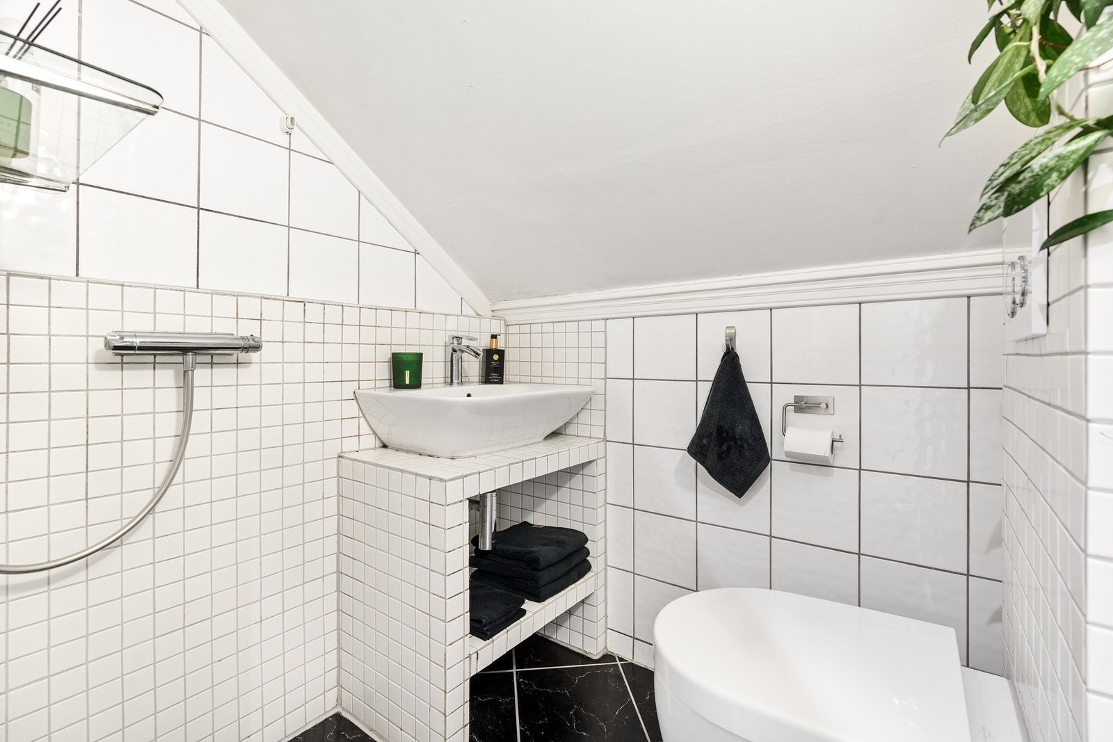 Badet har hvite fliser på veggene og sorte fliser på gulvet. Servanten er plassert på en flislagt hylle med oppbevaringsplass under. Vegghengt toalett og dusj med hånddusj er også installert. Galleribilde