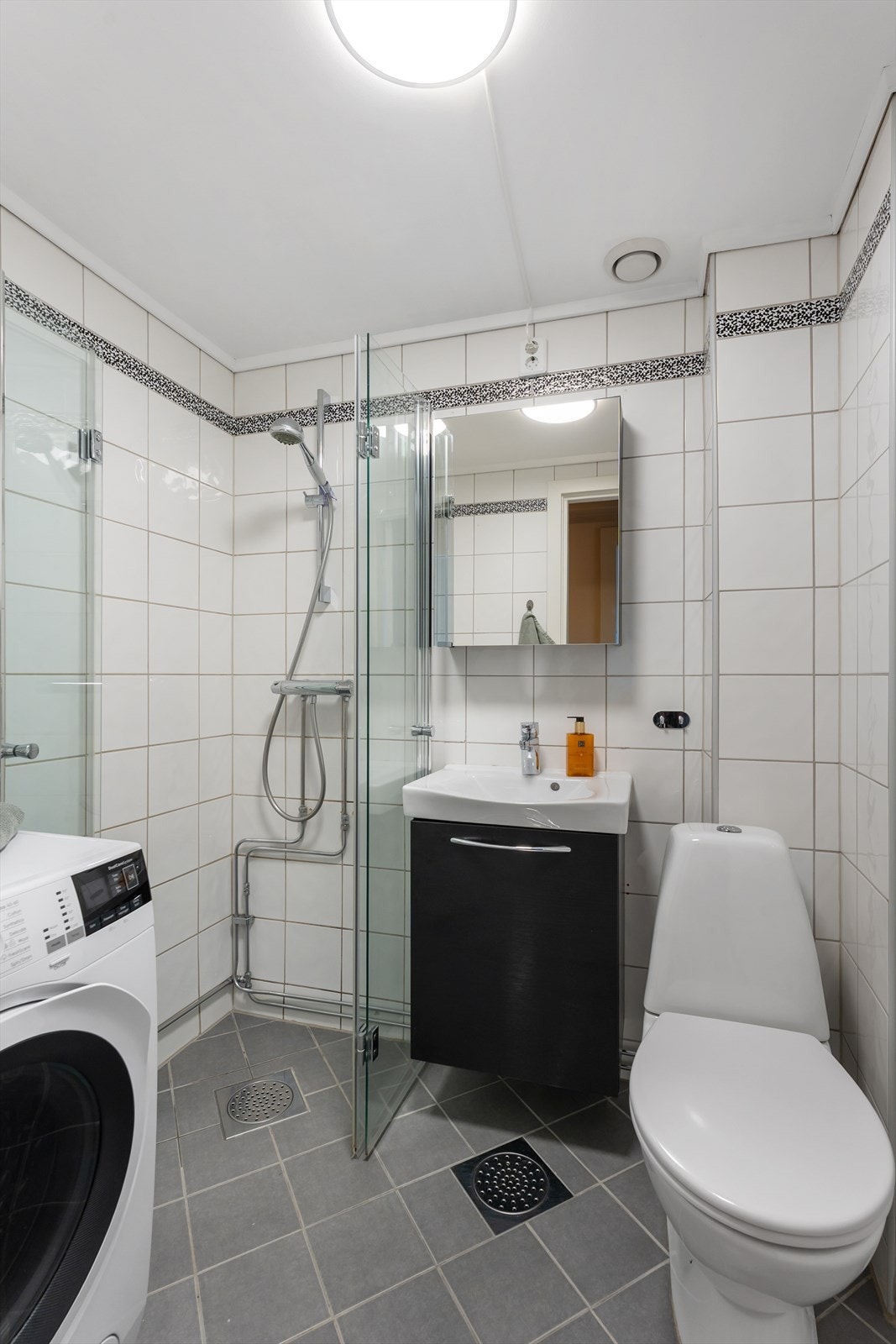 Videre er veggene flislagt og det er downlights i en slett himling, samt dusjhjørne med dusjdører i glass. Det er også opplegg for vaskemaskin og tørketrommel. Galleribilde
