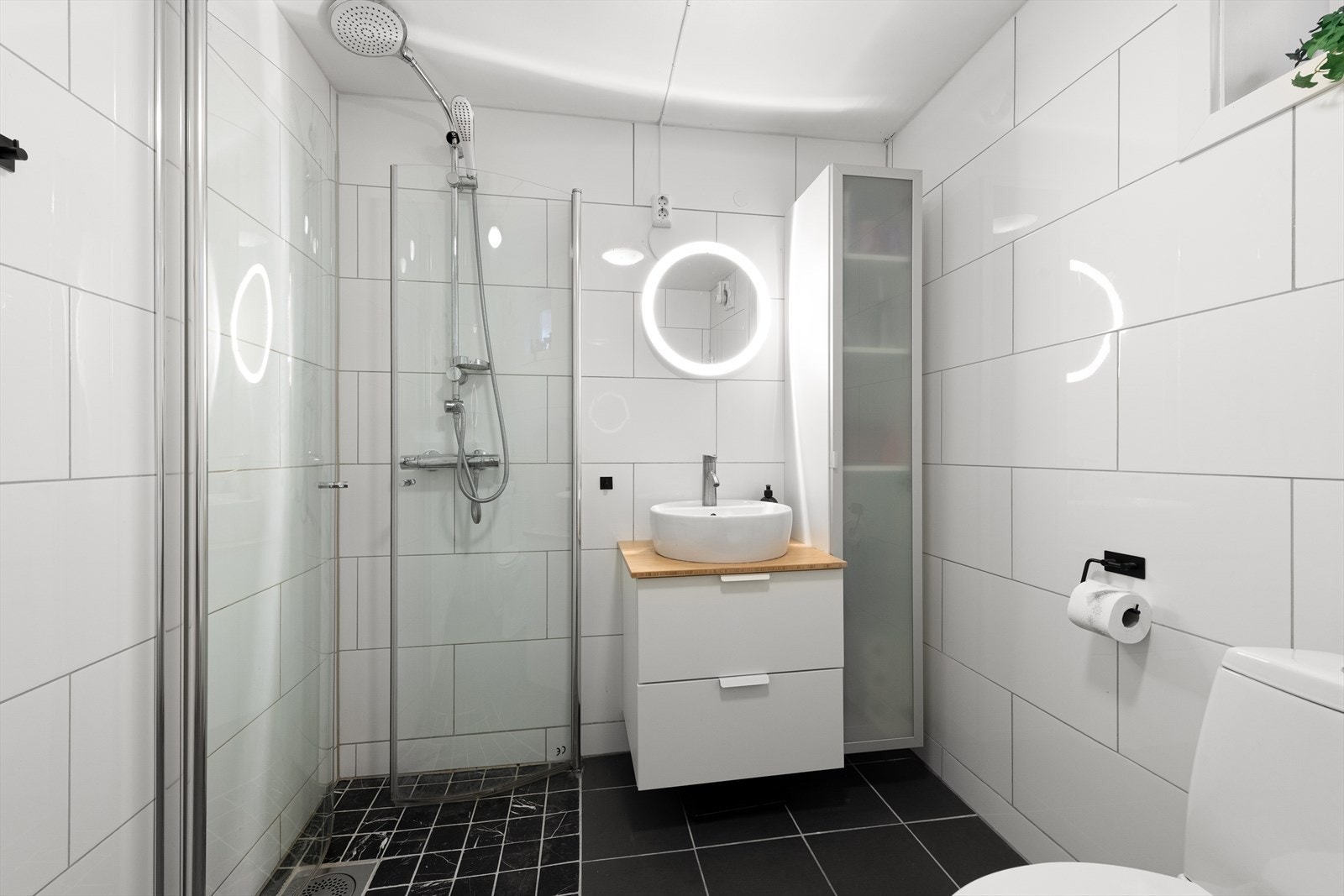 Badet er innredet med vegghengt toalett, innredning med servant, opplegg for vaskemaskin og dusjhjørne med glassdører. Galleribilde