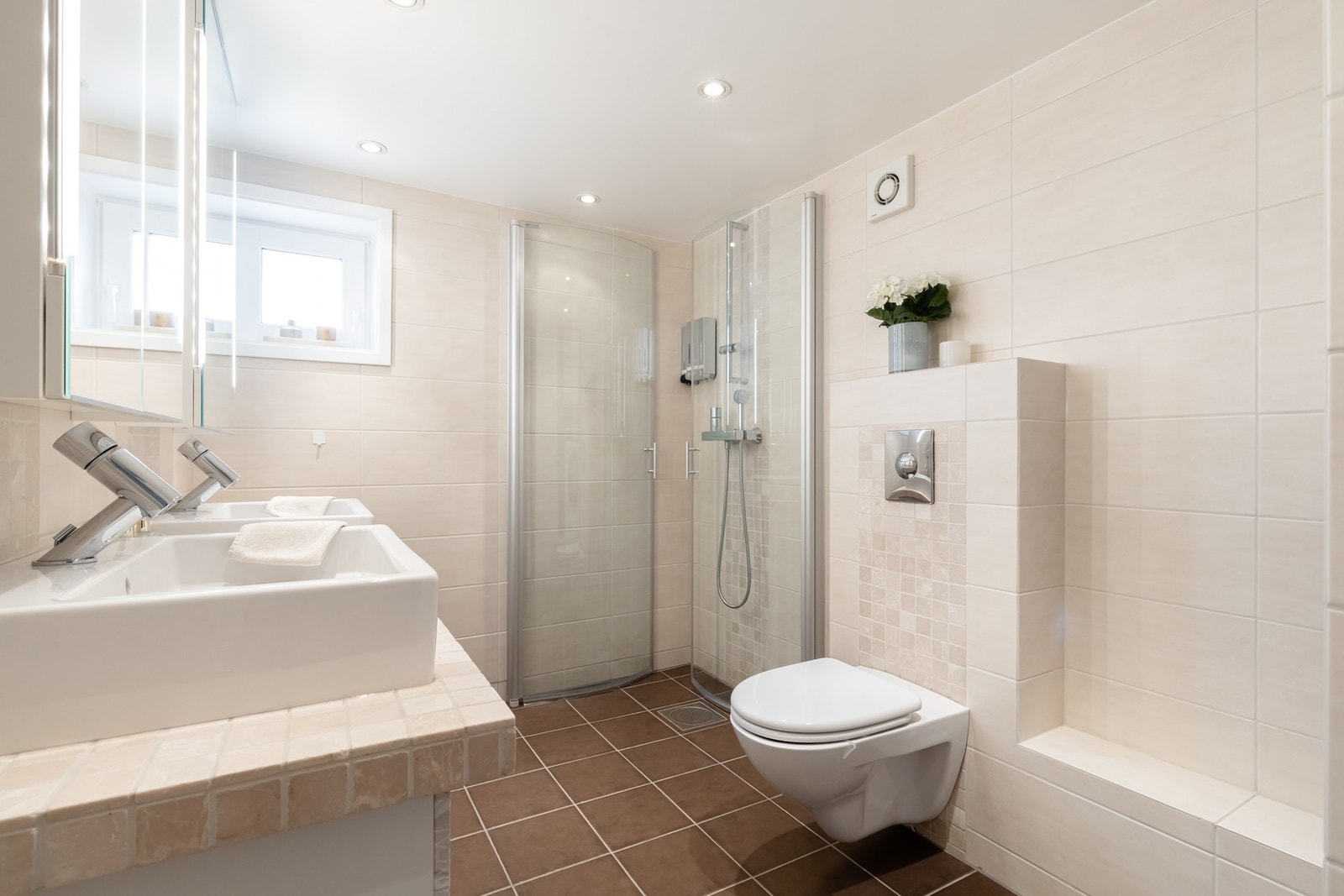 Badet er innredet med doble servanter på en flislagt benk med skuffer under, speil over hver servant, dusj med innadslående dører og vegghengt toalett. Galleribilde