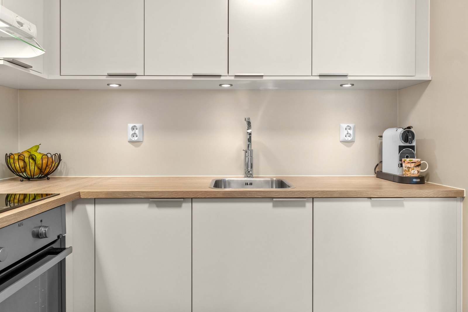 Glatte hvite fronter, laminat benkeplate med eik utførelse, nedfelt kum i rustfritt stål, krom blandebatteri samt moderne integrerte downlights under overskap. Galleribilde